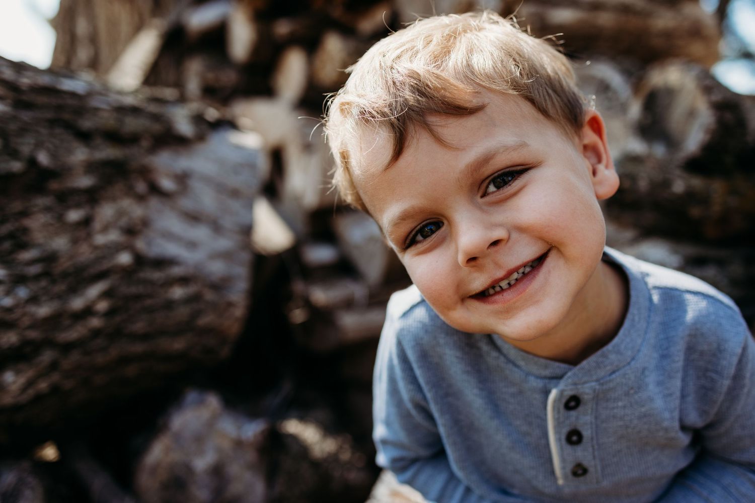 A young child wearing a gray henley shirt smiles brightly while sitting near wood logs outdoors.