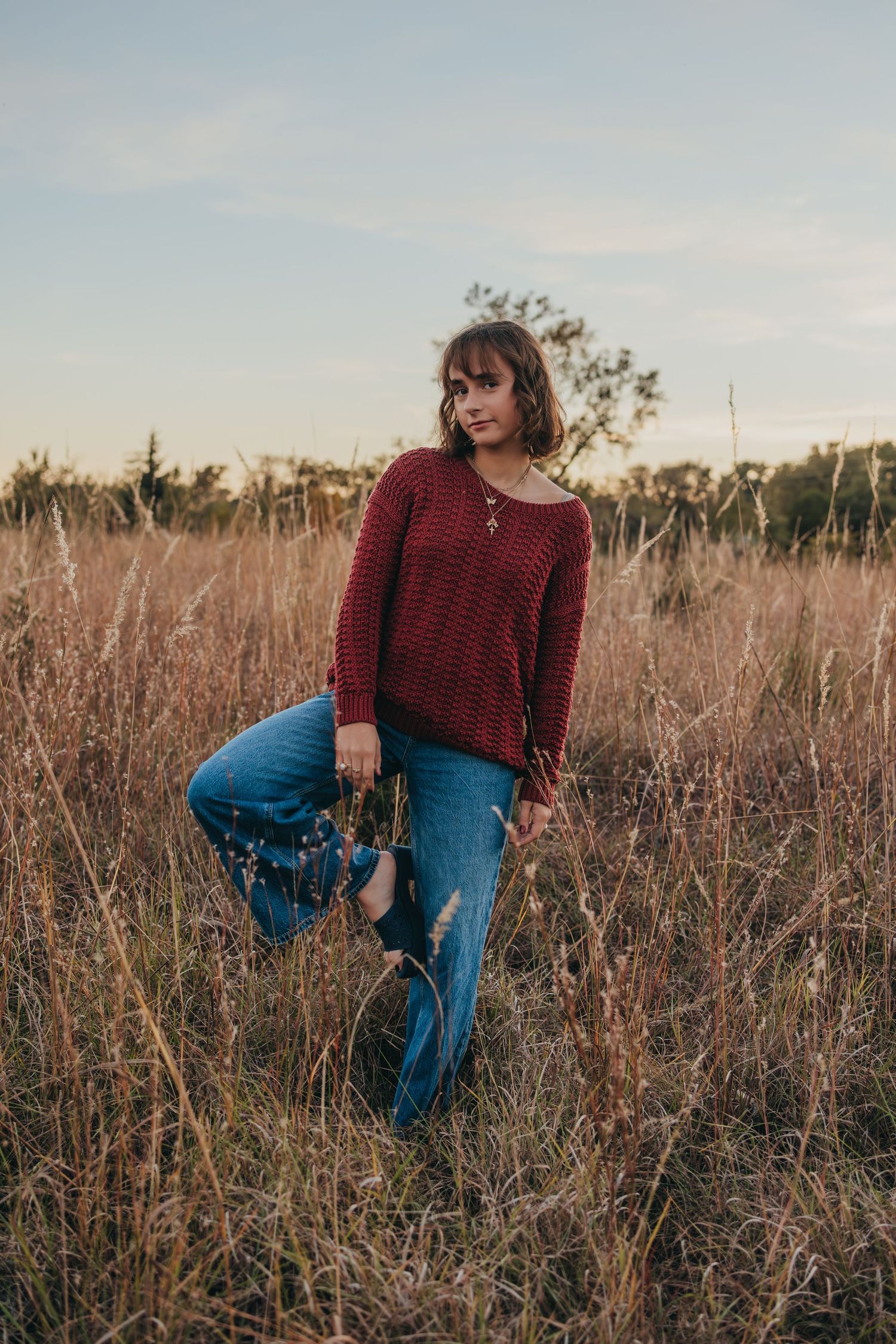 Young woman in red knit sweater and ripped jeans standing in golden field at sunset.