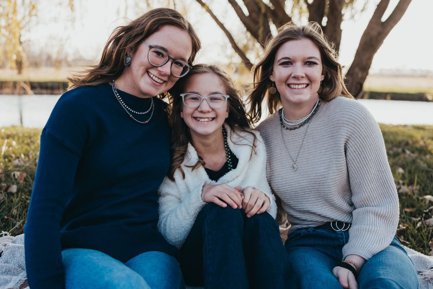Three people in cozy sweaters smile together outdoors on a sunny day.