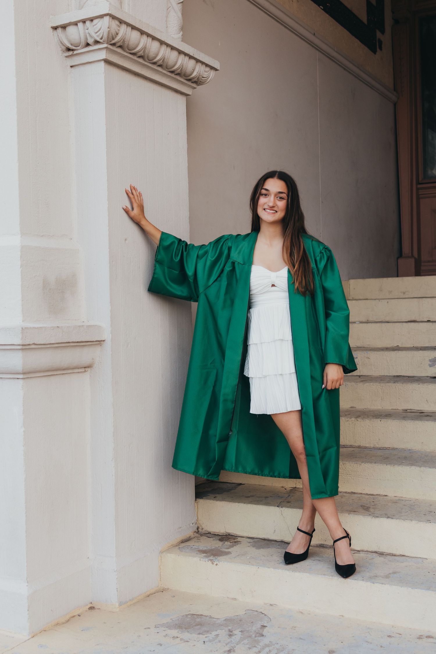 Young woman in green graduation gown and white dress poses on steps of academic building.
