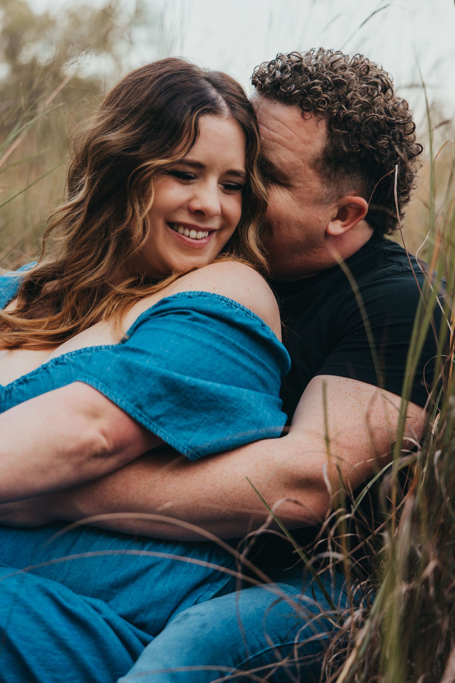 Happy couple cuddling in tall grass field, woman in blue off-shoulder dress smiling joyfully.