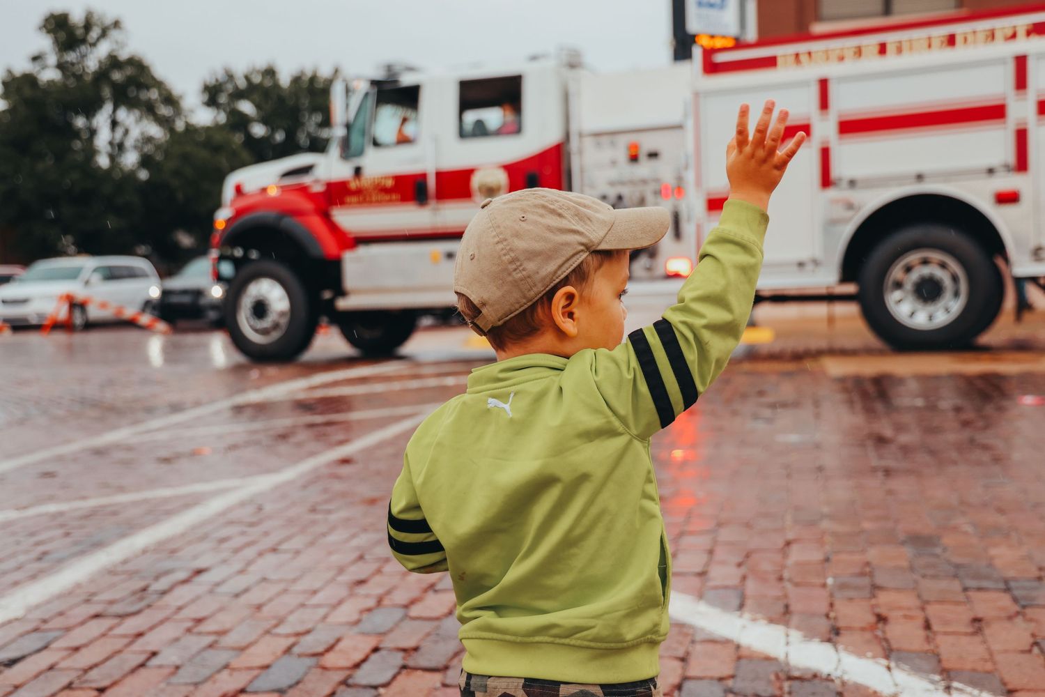 Young person in green jacket waving at passing fire trucks on rainy street.