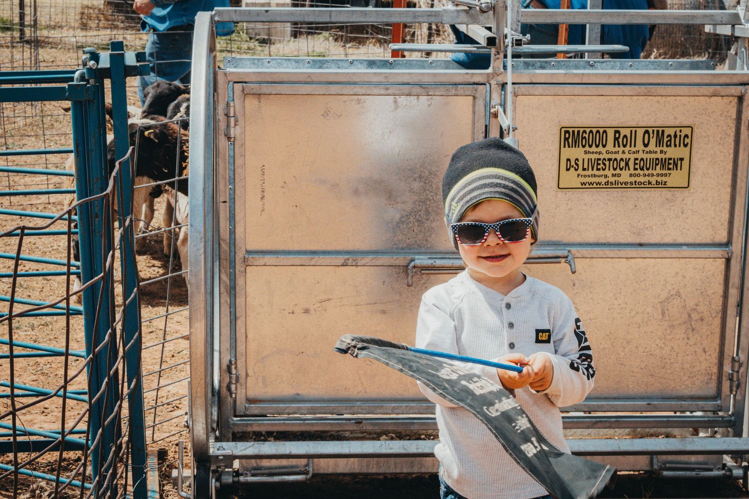 Young rancher with sunglasses and beanie holds clipboard by livestock handling equipment.