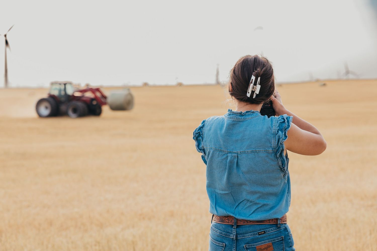 Person wearing blue shirt and jeans watching a red tractor in a vast wheat field.