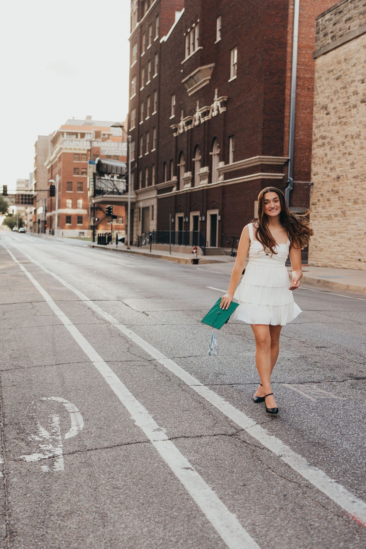 Young woman in white dress holding green clutch walks urban street past brick buildings.