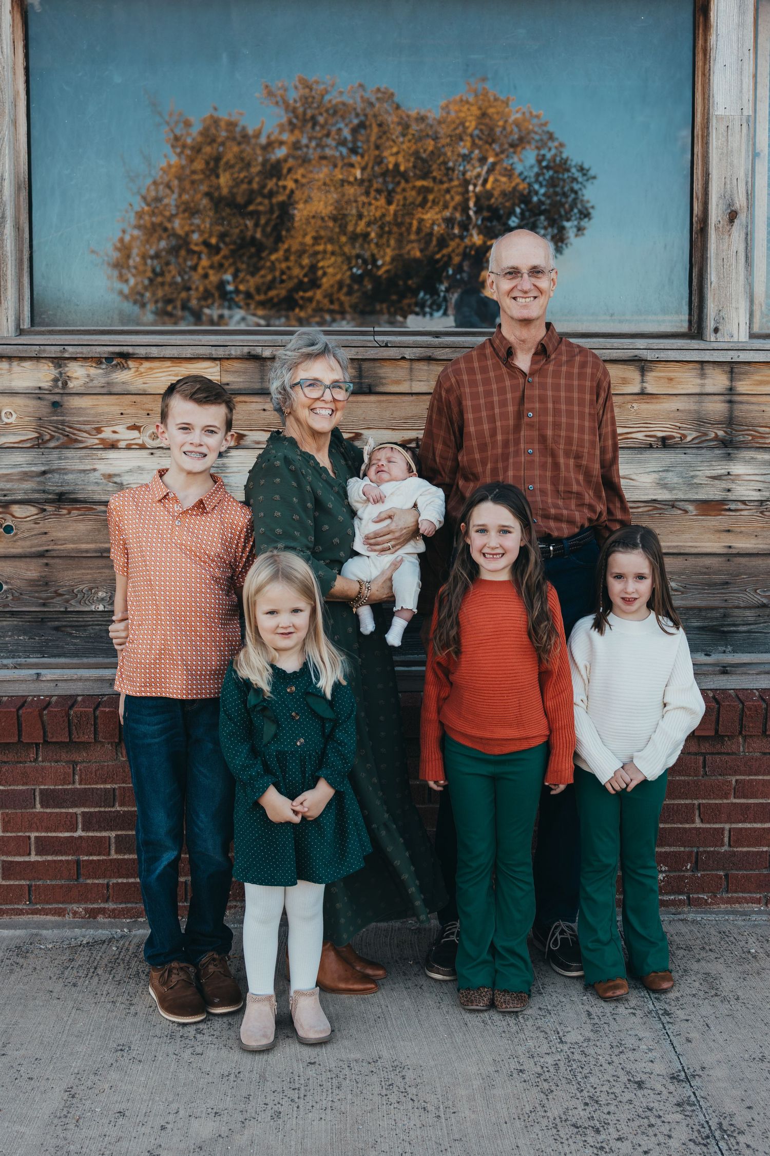 Multi-generational family portrait with grandparents, four children, and a newborn baby posing outdoors near a rustic brick wall.