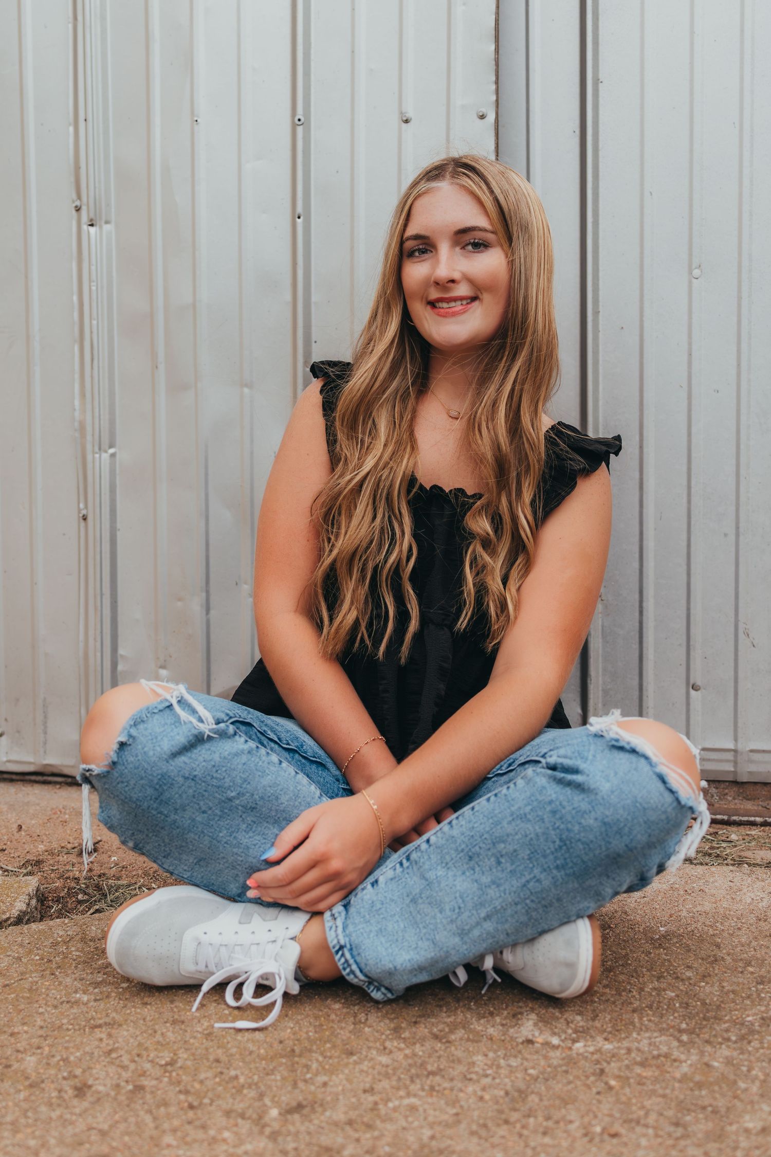A person wearing a black top and light jeans sits casually on the ground in a series of candid poses against a metal wall.