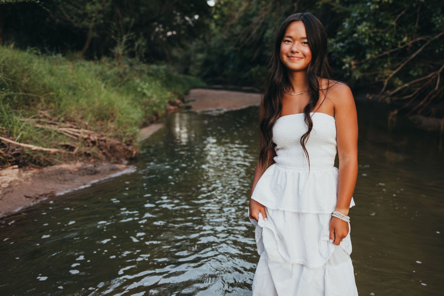 Woman in flowing white sundress smiles while standing by a peaceful creek on a summer day.
