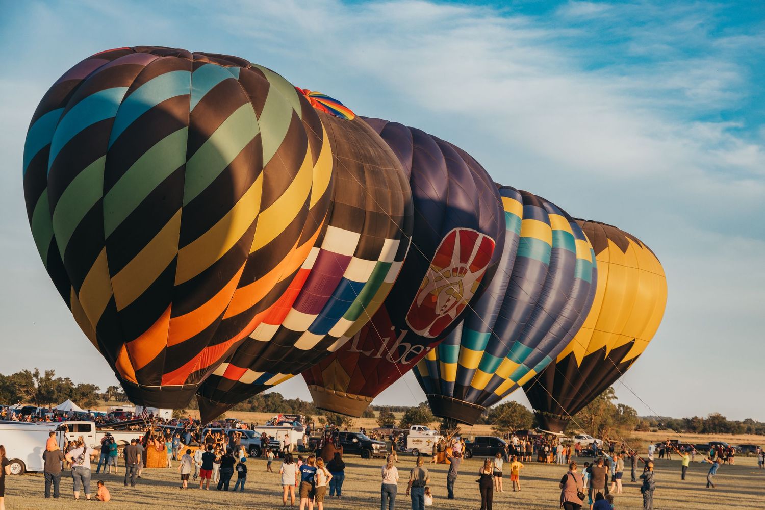 Colorful hot air balloons line up on a field with spectators gathering around.