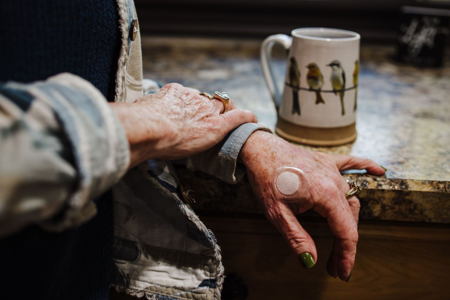 Close-up shot of a weathered hand resting on a table beside a ceramic mug decorated with bird designs.