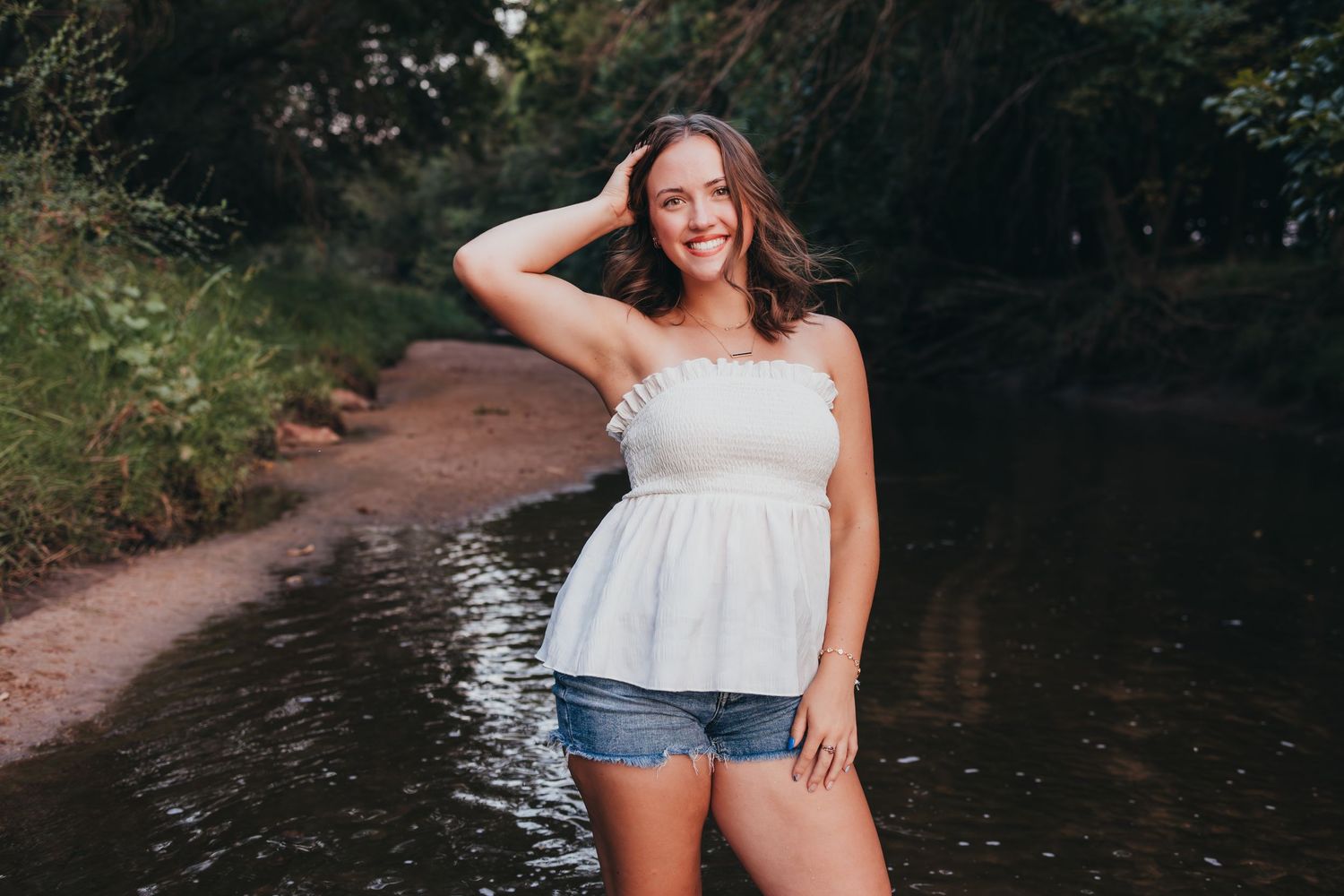 Smiling woman in white top and denim shorts posing along a scenic wooded path by a stream in summer.