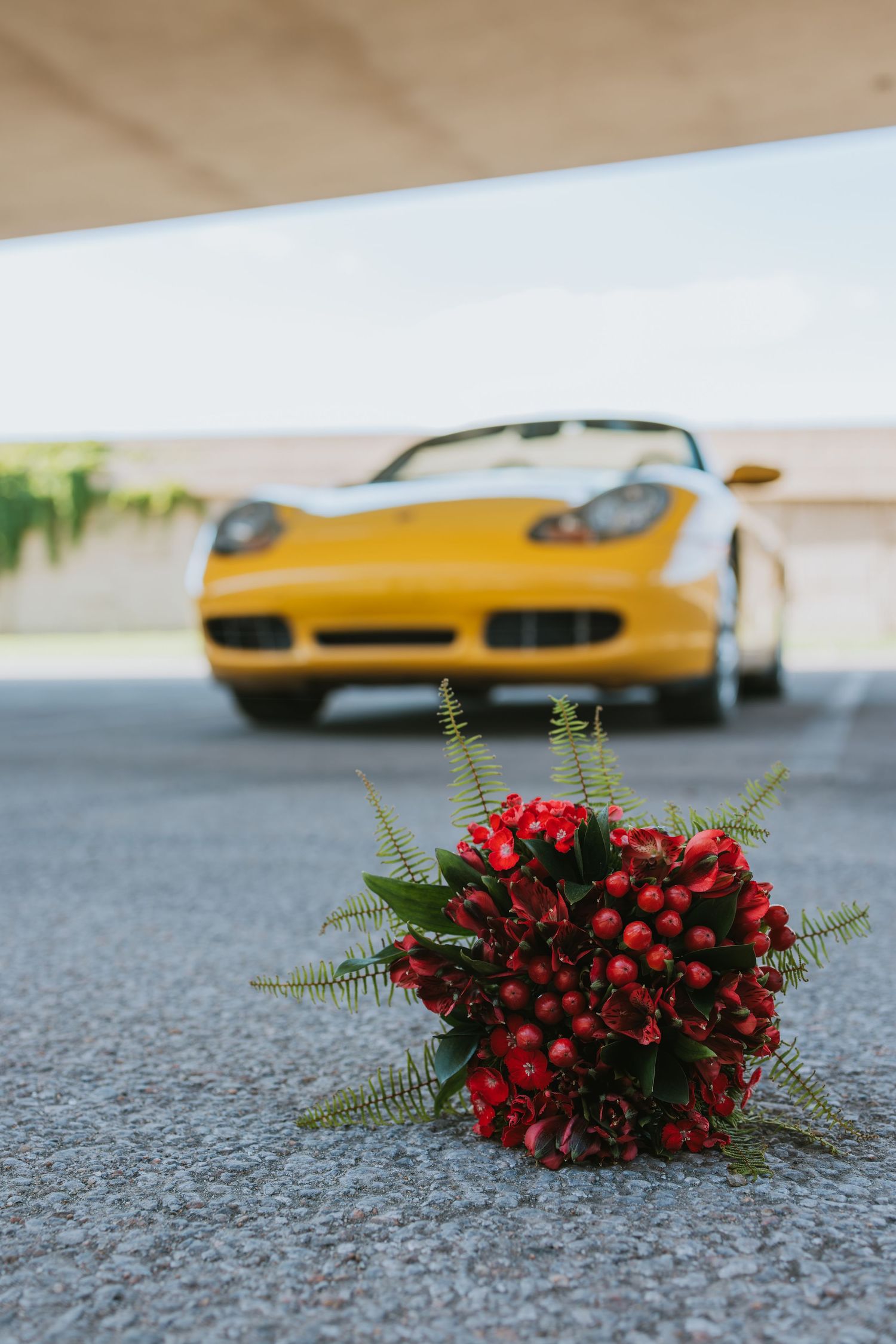 A red floral bouquet with ferns sits on pavement with a yellow Porsche sports car blurred in the background.