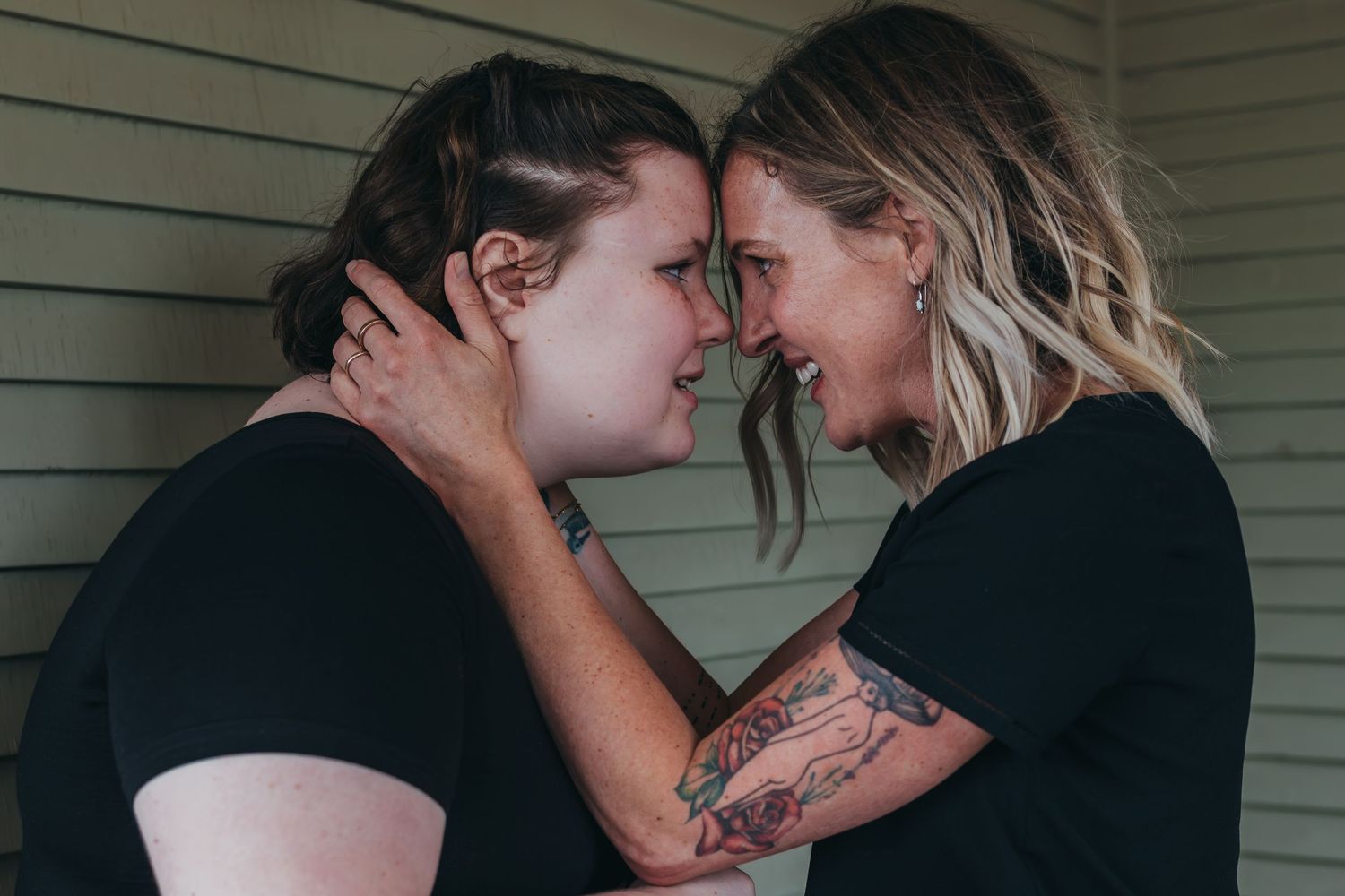 Two people in black shirts share an intimate moment together, with one person displaying a visible arm tattoo.