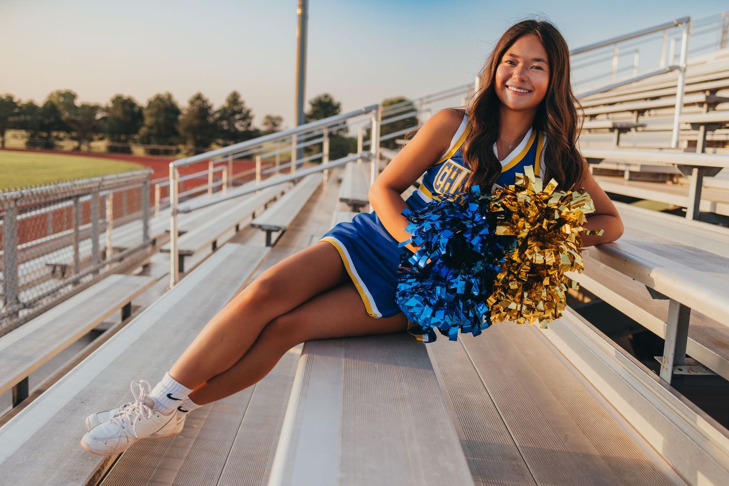 A cheerleader with pom poms poses on bleachers during a golden hour photoshoot in an outdoor stadium.