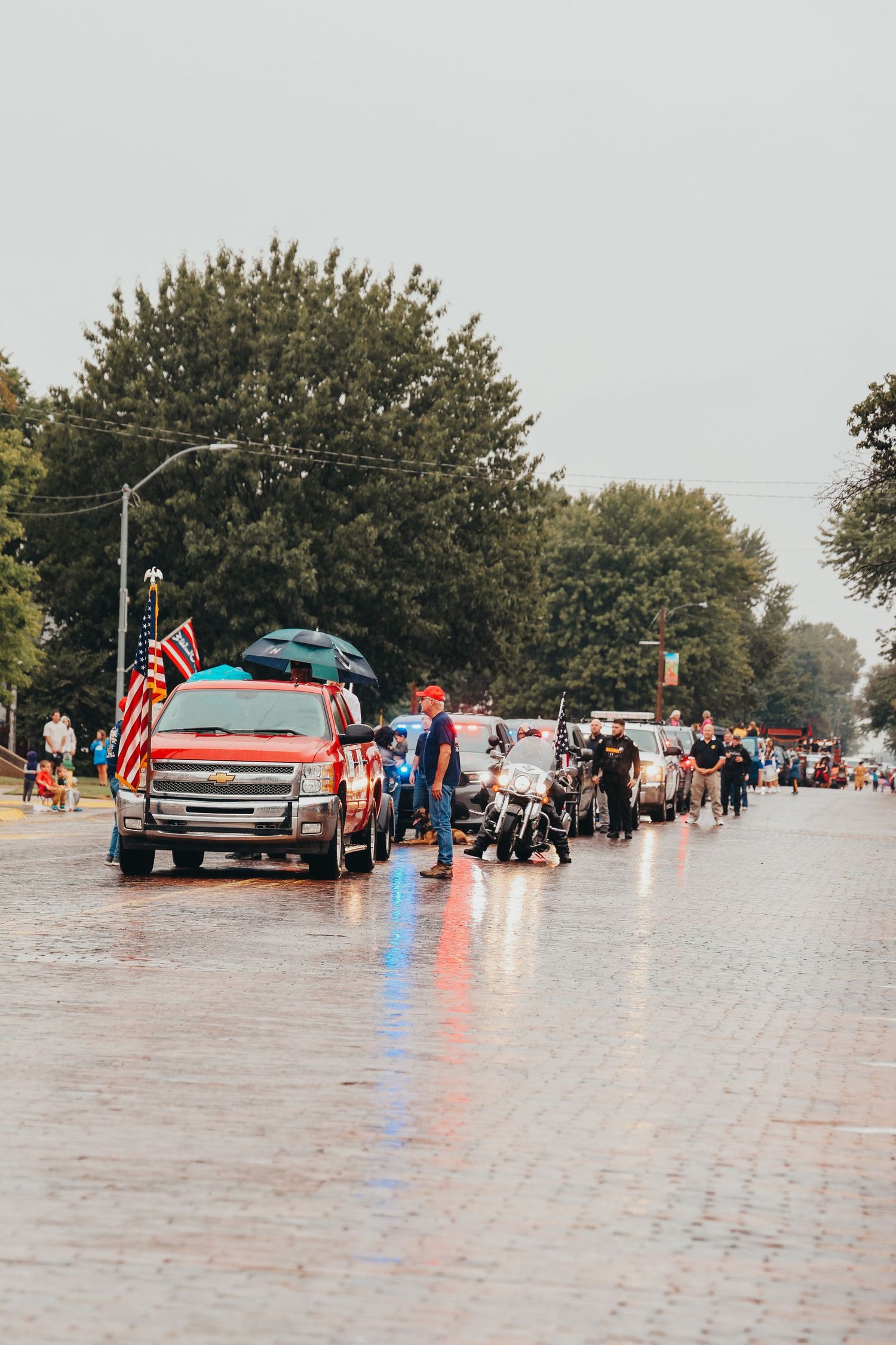 A crowd gathers along a wet street for a parade with American flags and trucks.