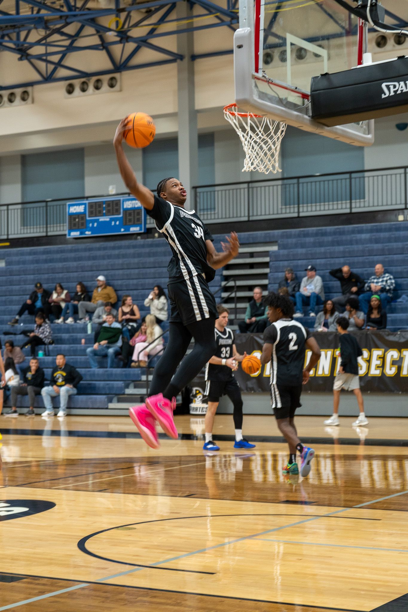 Basketball player in black uniform performs a layup during a game in a gymnasium with spectators in the bleachers.
