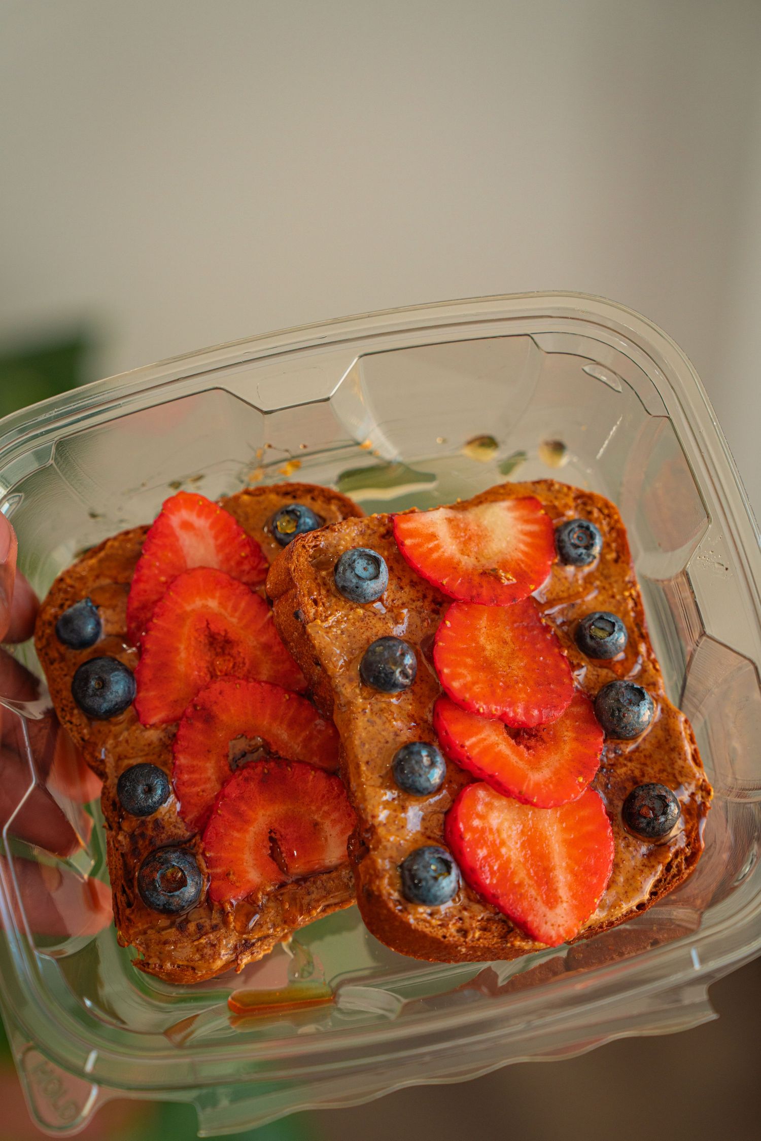 Fresh toast topped with sliced strawberries and blueberries in a clear plastic container.