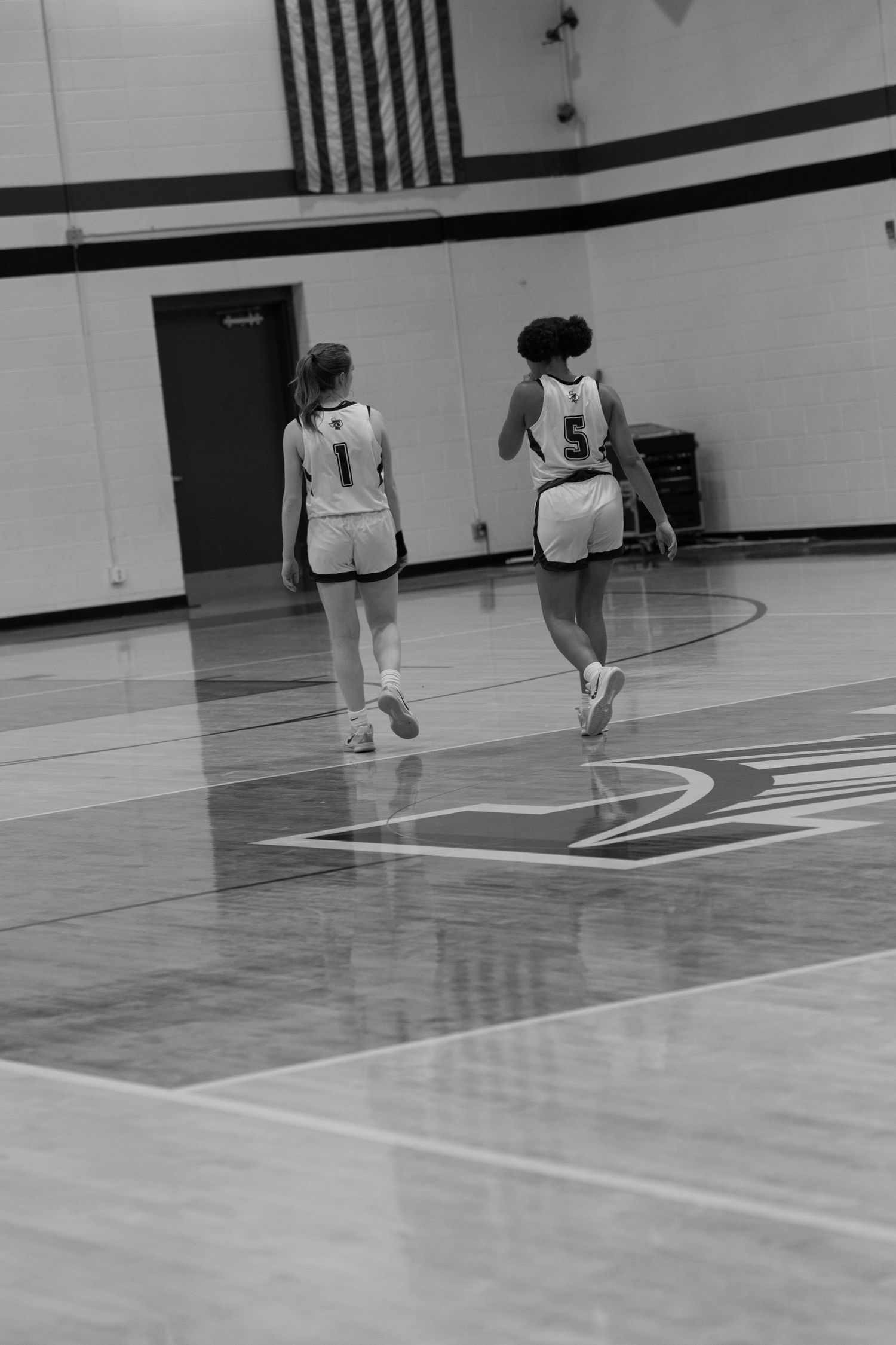Two basketball players in white jerseys numbered 1 and 5 walk across a gymnasium court during a game.