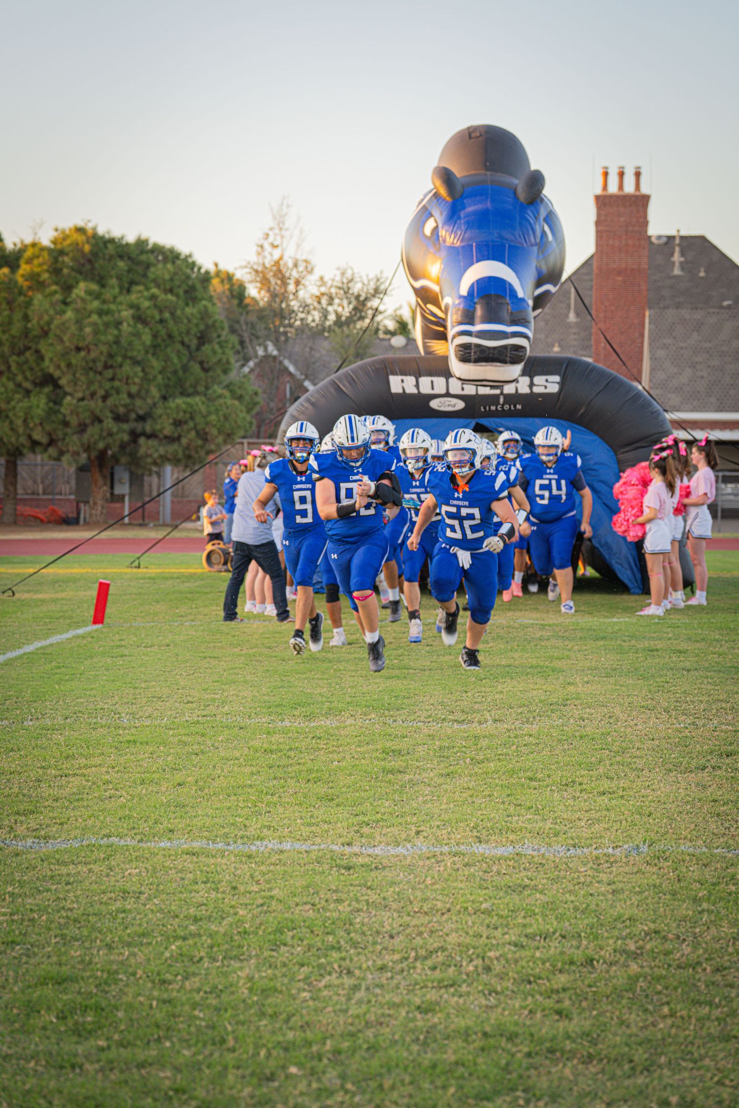 High school football team in blue uniforms runs onto field through inflatable helmet tunnel during game entrance.