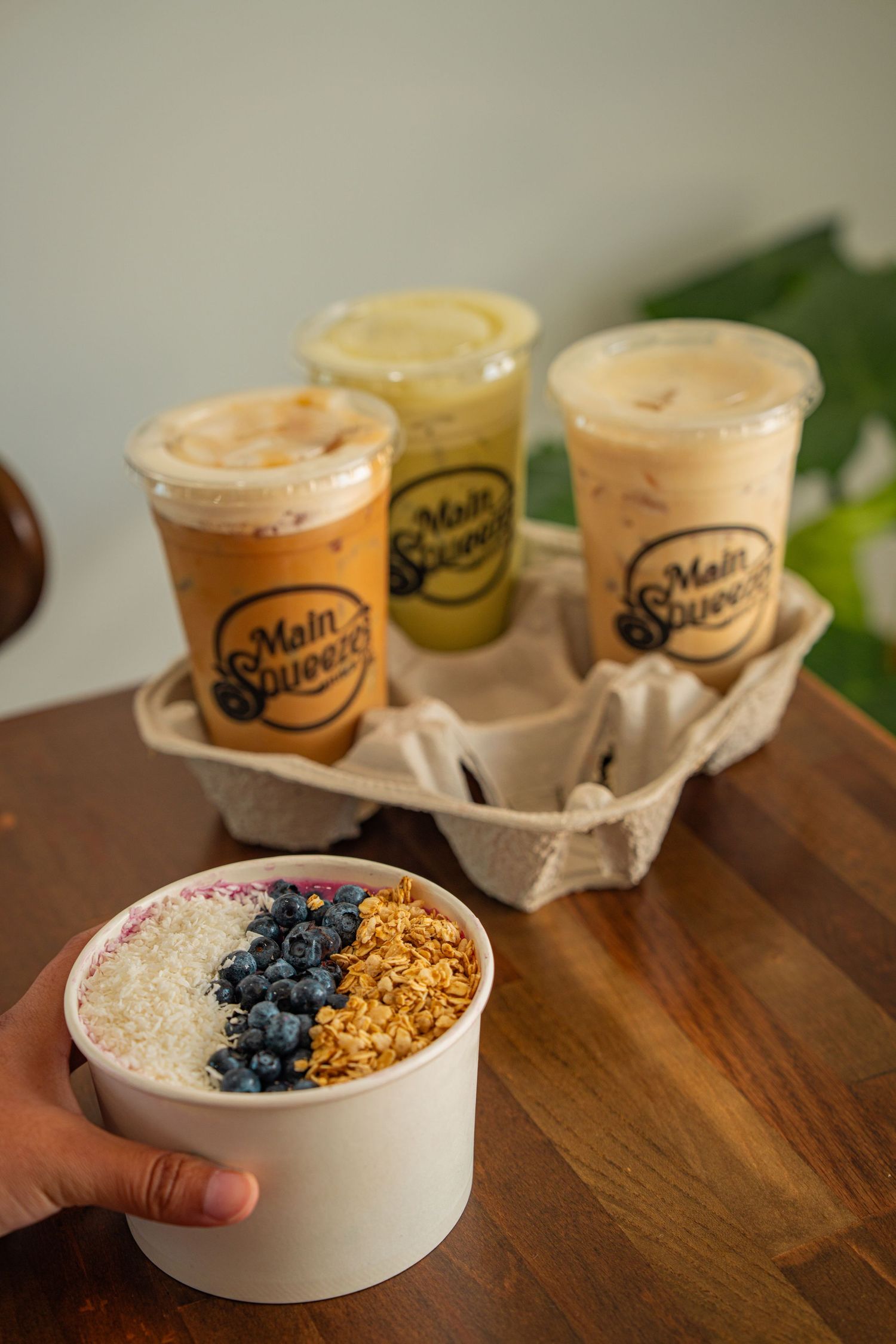 Takeout cups of smoothies arranged on wooden table with blueberries and granola in a ceramic bowl.