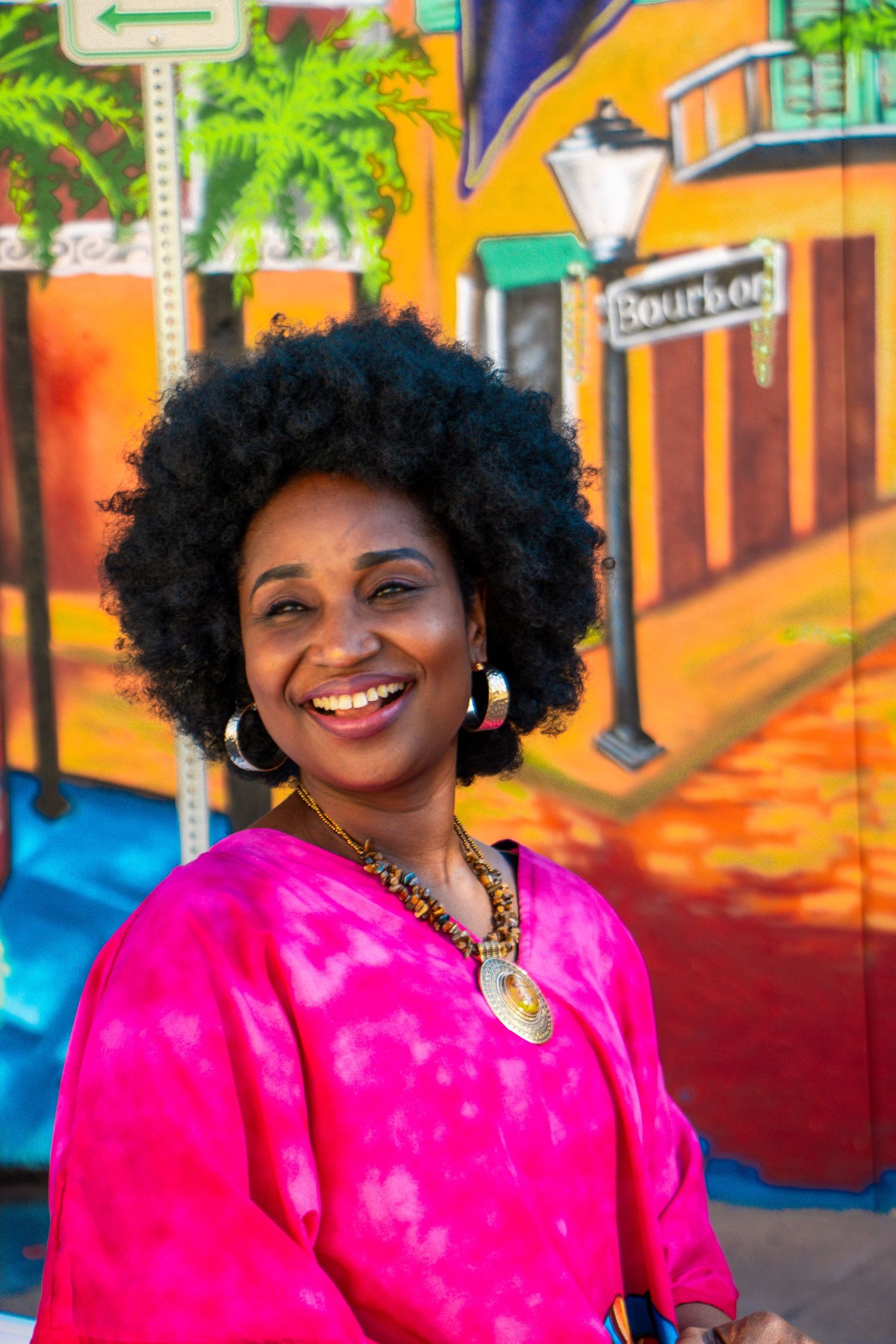A person in vibrant pink attire smiling against a colorful mural background.
