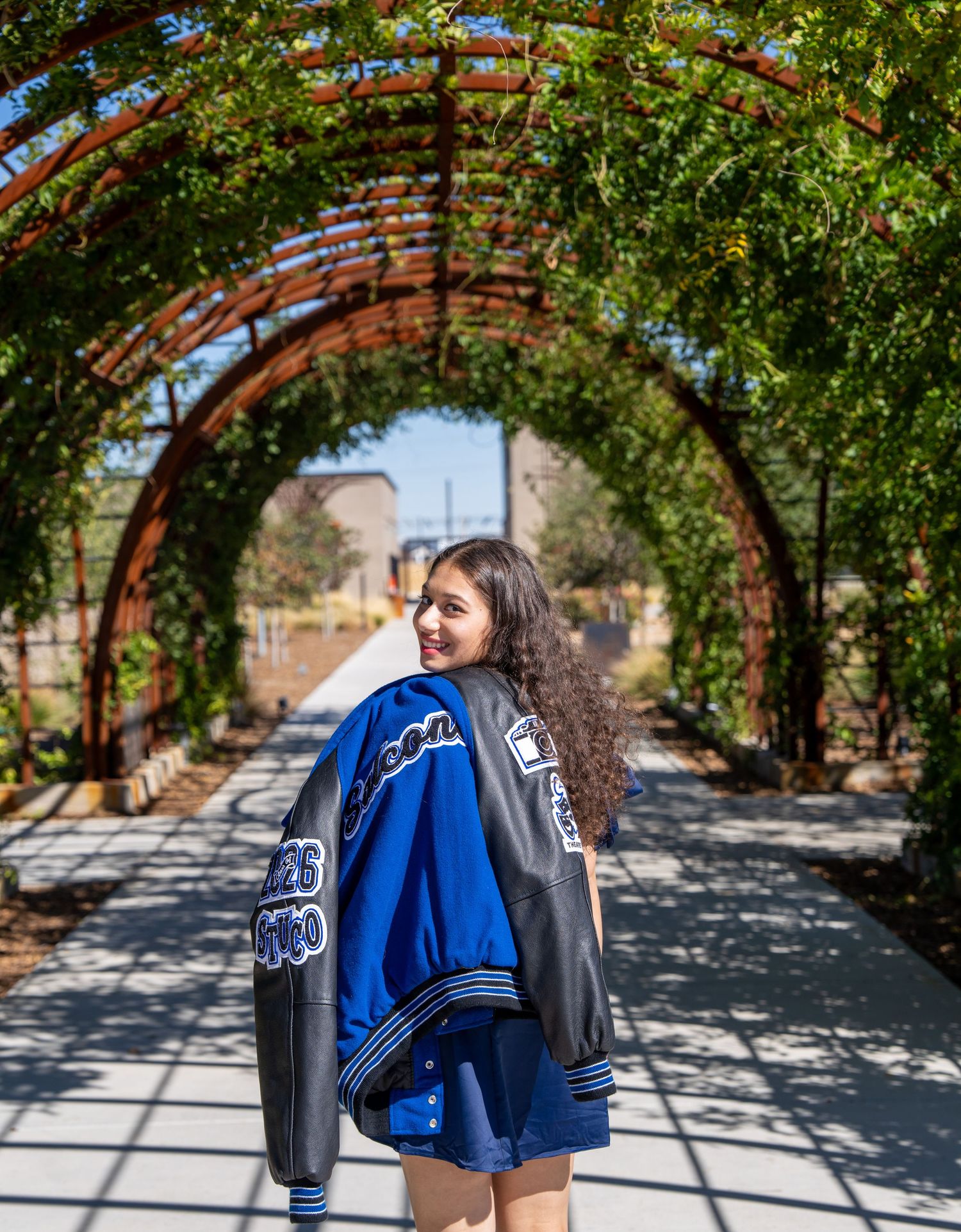 A person in a blue varsity jacket poses under a vine-covered archway on a sunny path.
