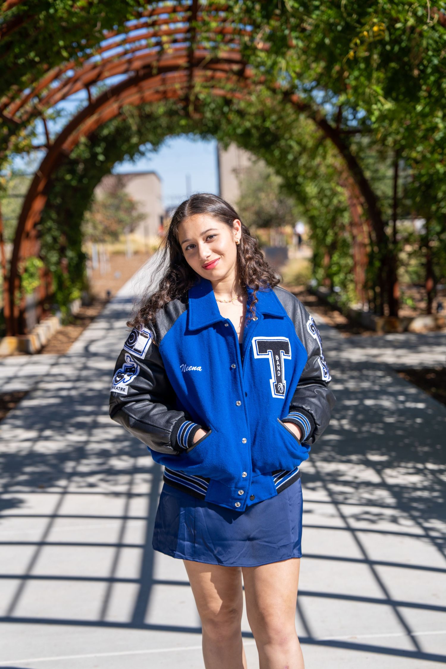 Student wearing a blue and black varsity letterman jacket poses under an arched garden trellis on a sunny day.