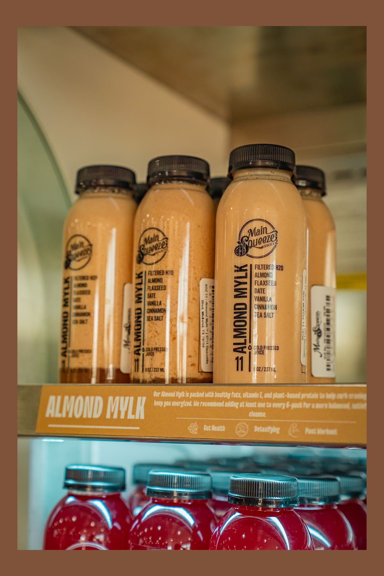 Rows of bottled beverages including orange-colored almond mylk drinks and red juices arranged on refrigerator shelves.