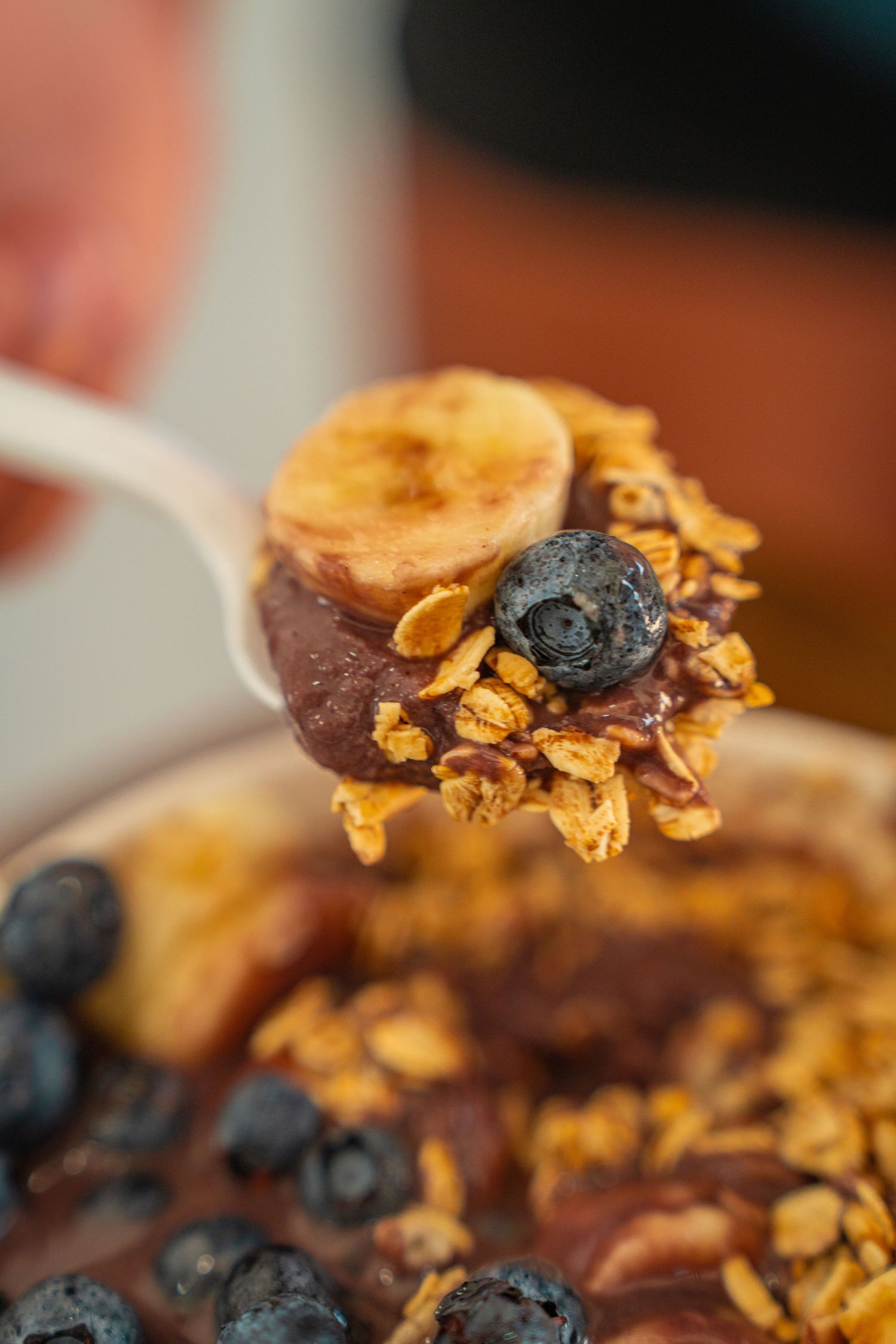 A spoonful of acai bowl topped with granola, banana slices and fresh blueberries against a blurred background.