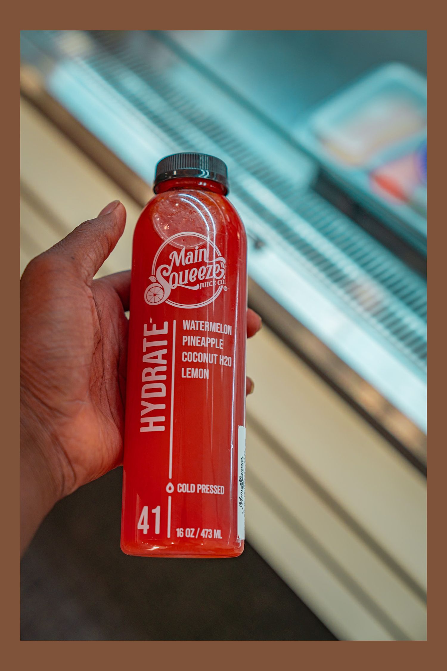 Hand holds a red watermelon pineapple coconut cold pressed juice beverage can against a blurred background.