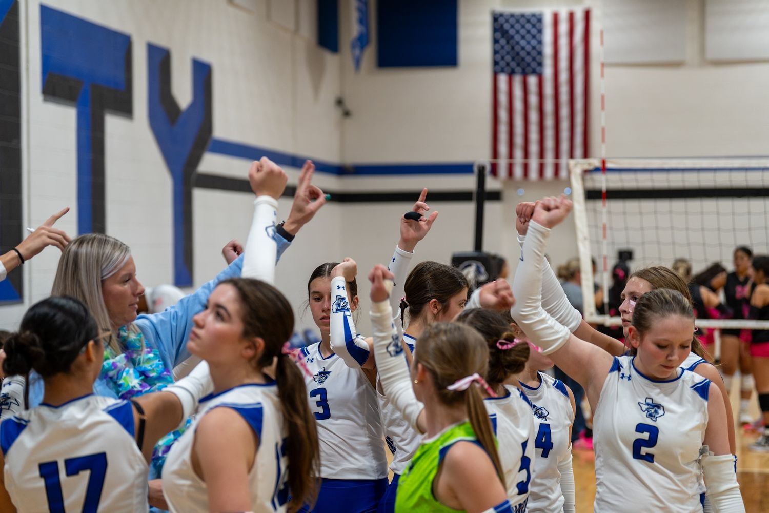 High school volleyball players celebrate a point during an indoor match with American flags visible in the gymnasium.