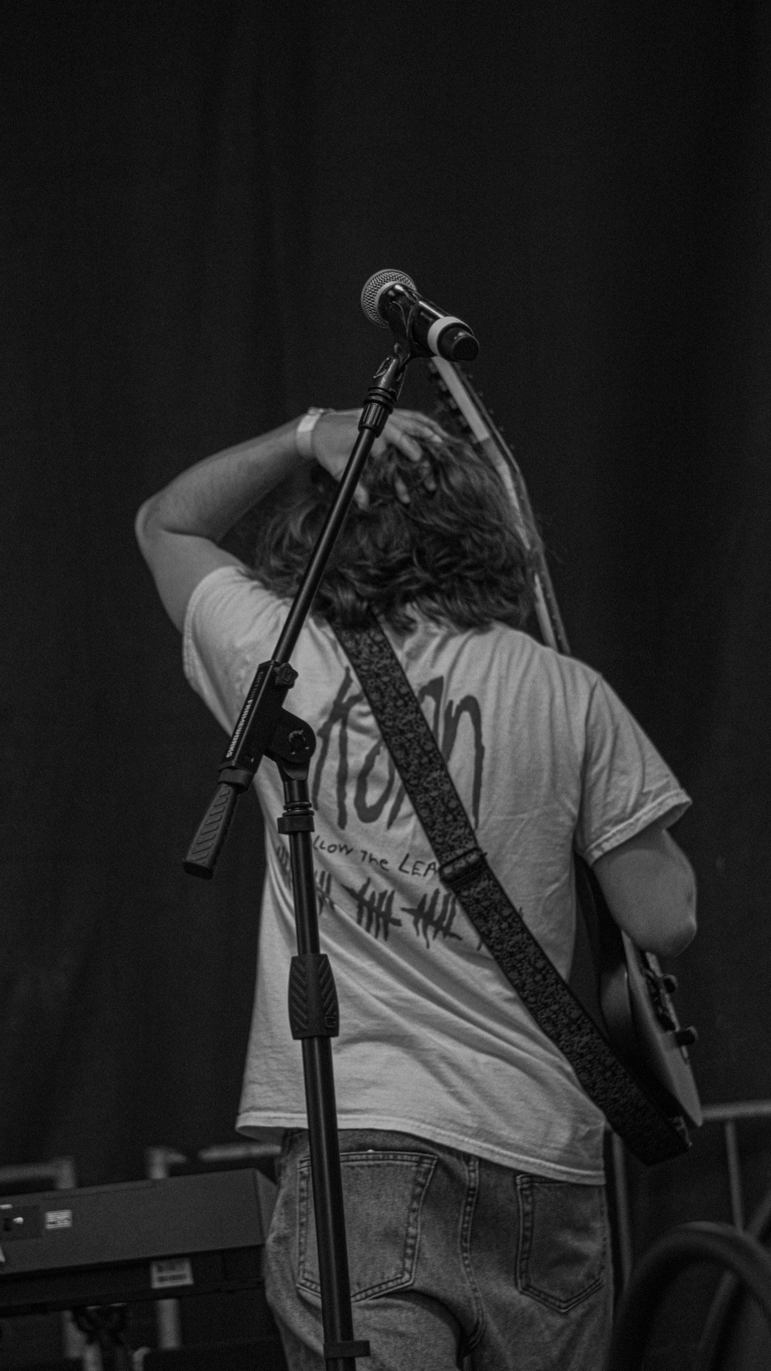 Musician performing on stage seen from behind with microphone stand and guitar strap in black and white.