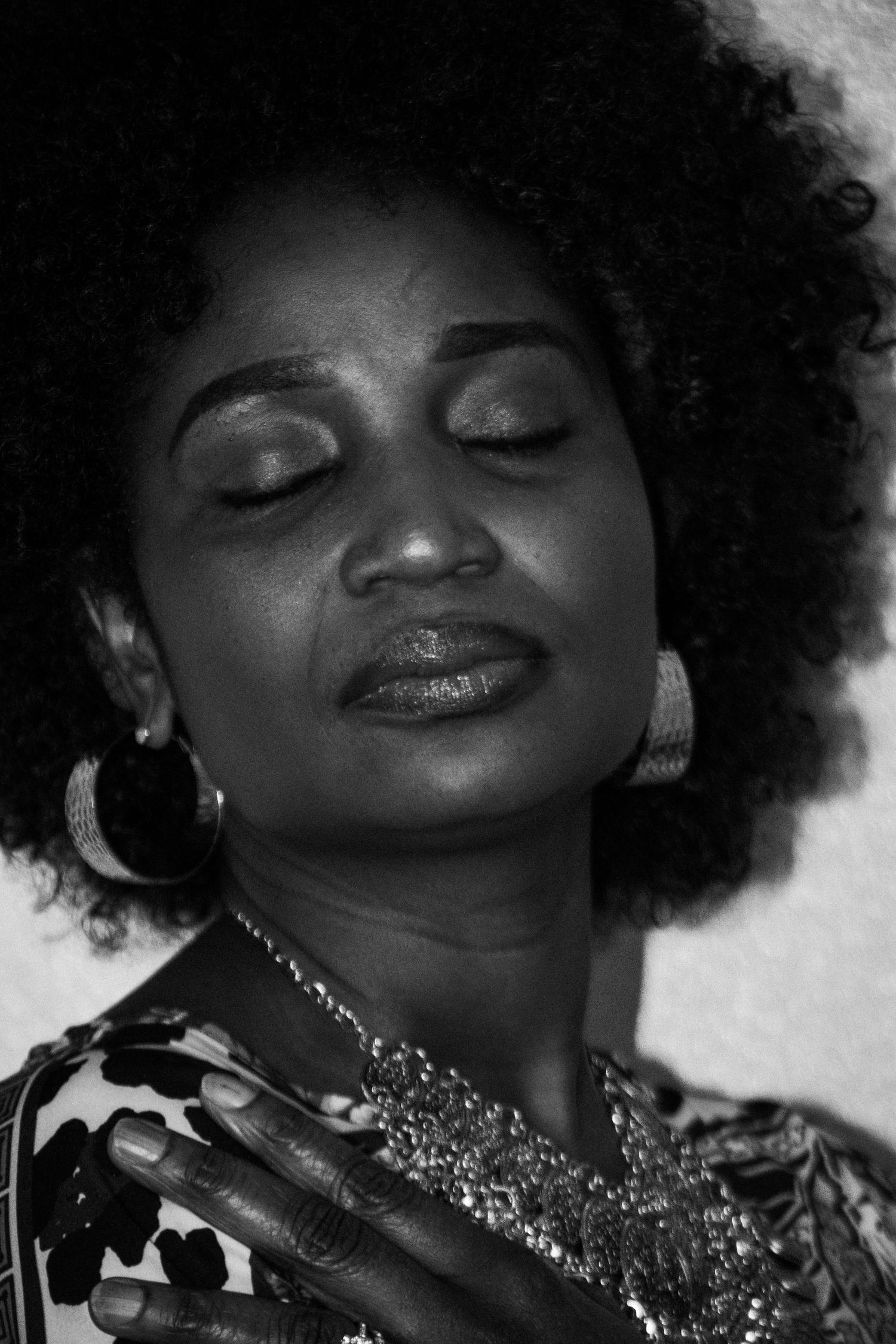 An artistic black and white portrait featuring natural hair, closed eyes, and statement jewelry.