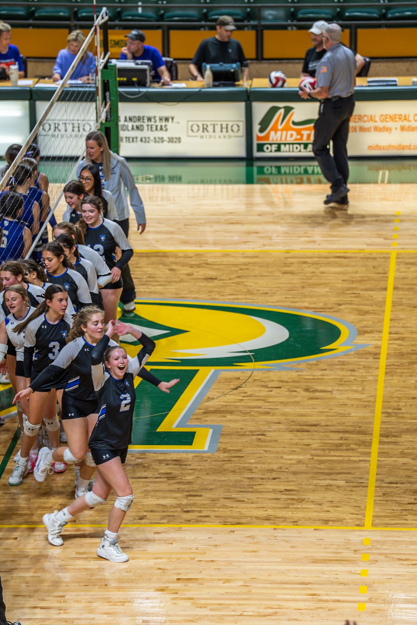 Athletes in dark jerseys leap and dive during an intense volleyball match on a gymnasium court.