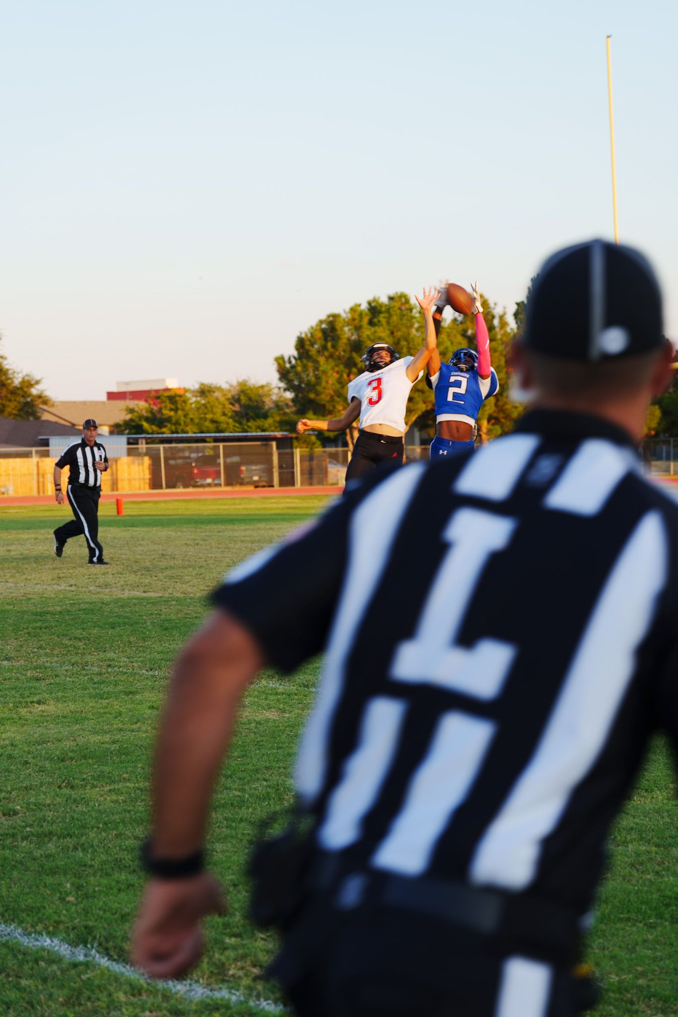 Football referees in black and white striped uniforms stand on a grassy field during a game at sunset.