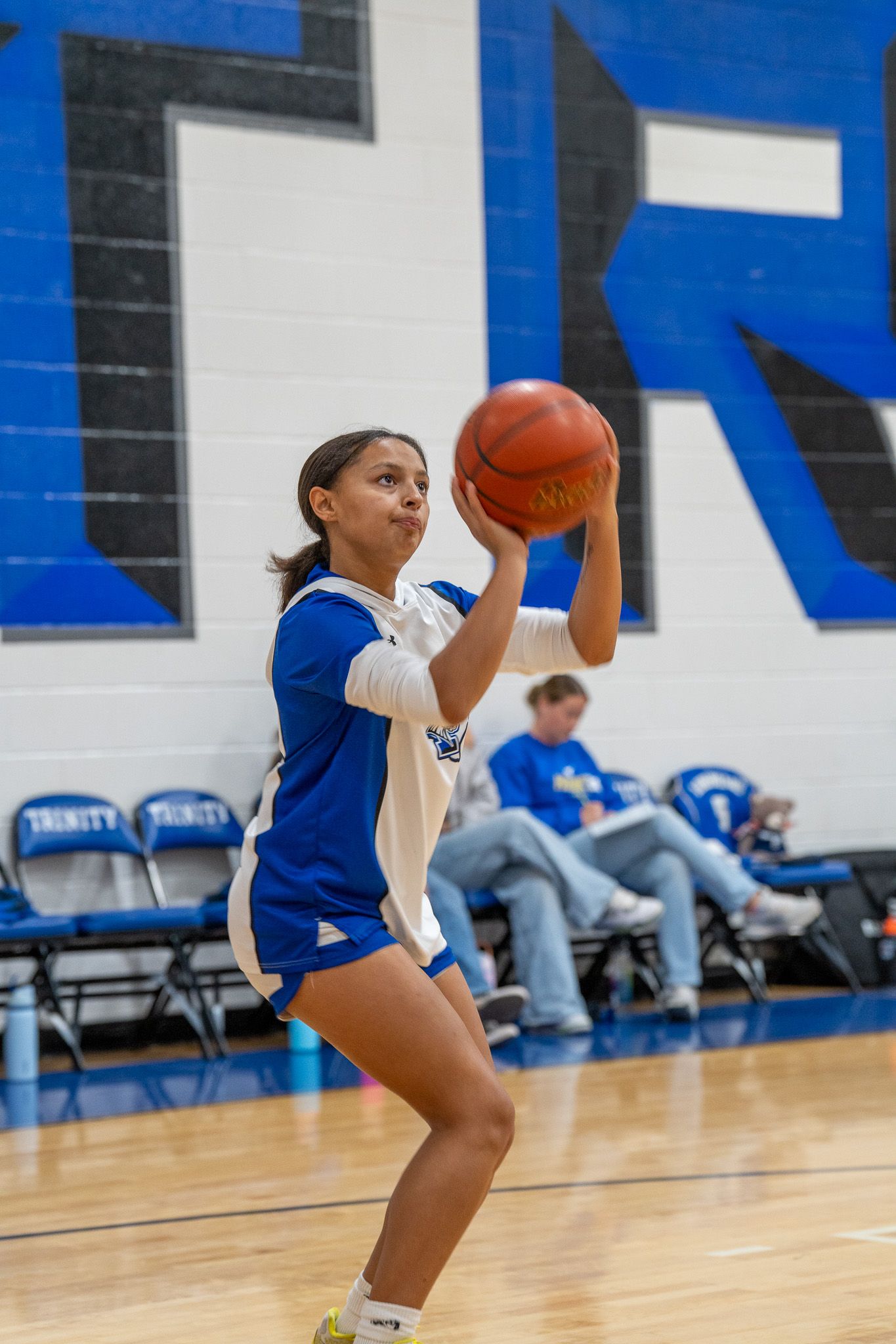 Female basketball player in blue uniform crouches low to catch a pass during practice in a gymnasium.
