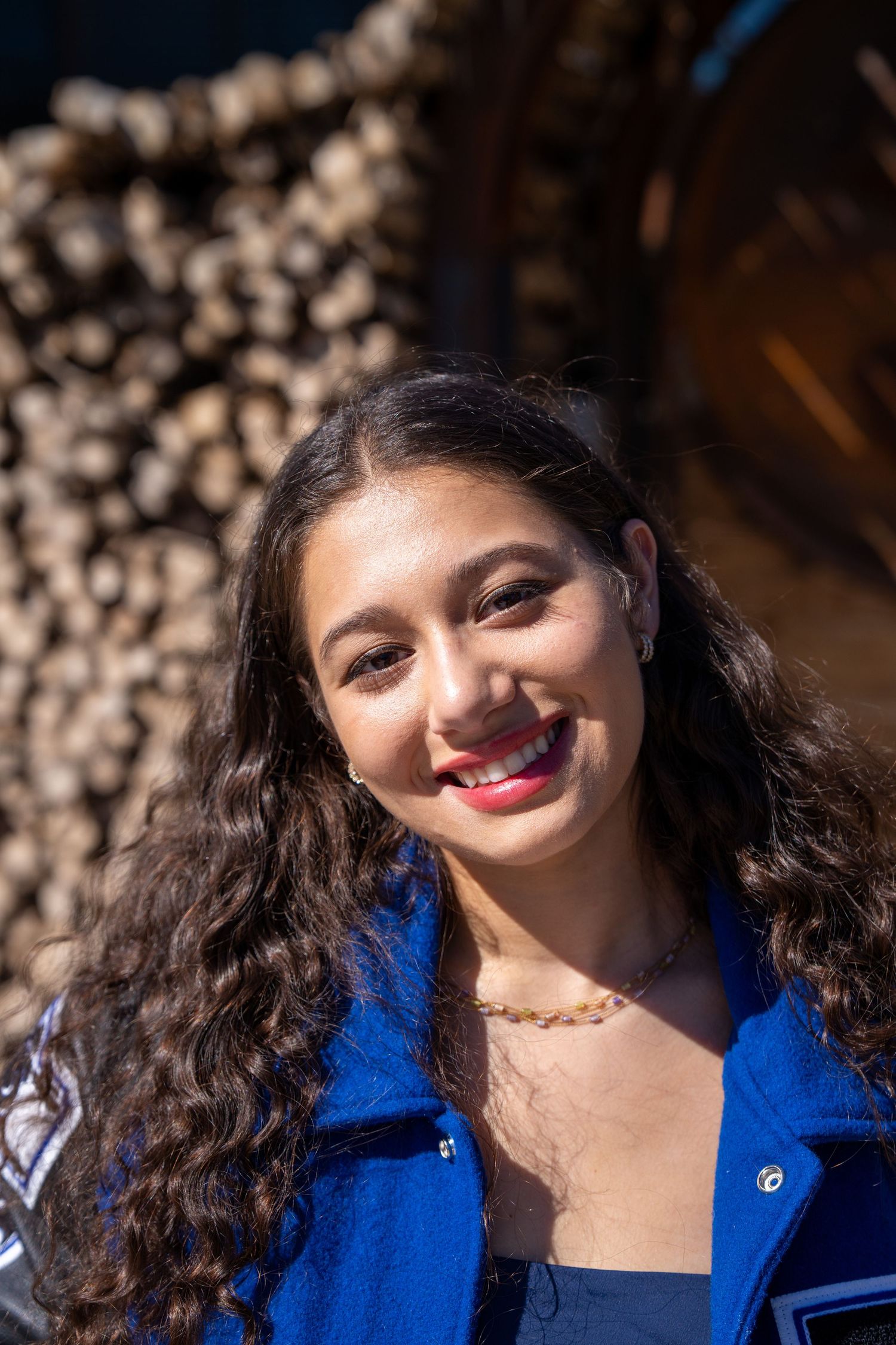 A woman in a bright blue jacket smiles warmly while posing outdoors with blurred cherry blossoms in the background.