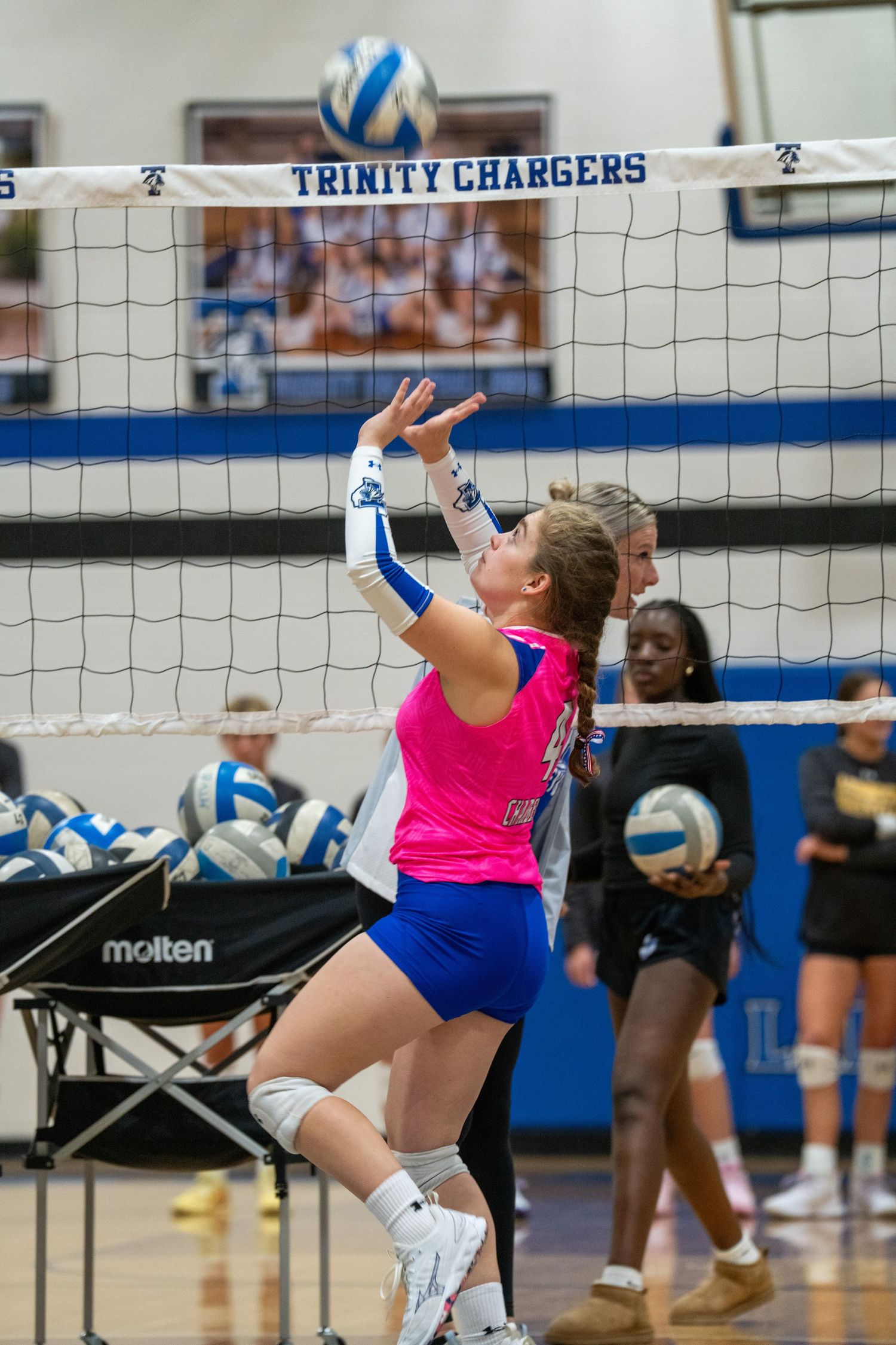 Indoor volleyball players compete at the net during an intense match at Trinity Chargers gymnasium.