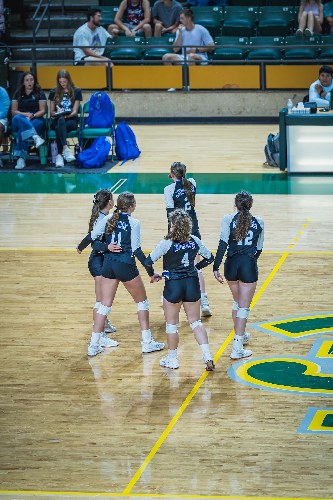 Volleyball players in navy uniforms huddle together on a wooden court with yellow and green markings.