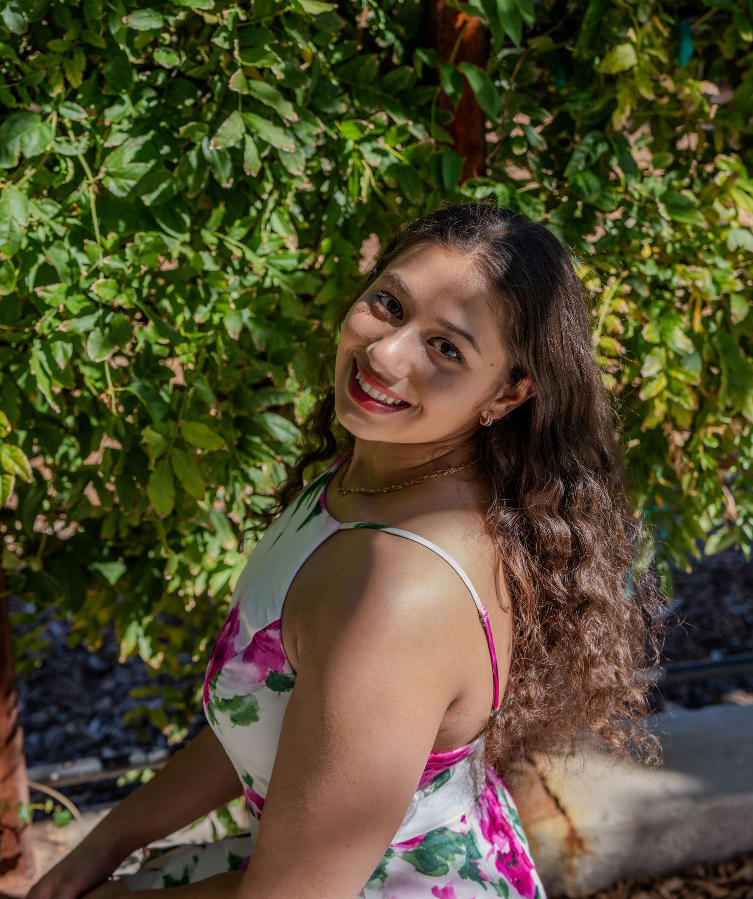 A smiling person with long wavy hair poses outdoors against lush green foliage on a sunny day.