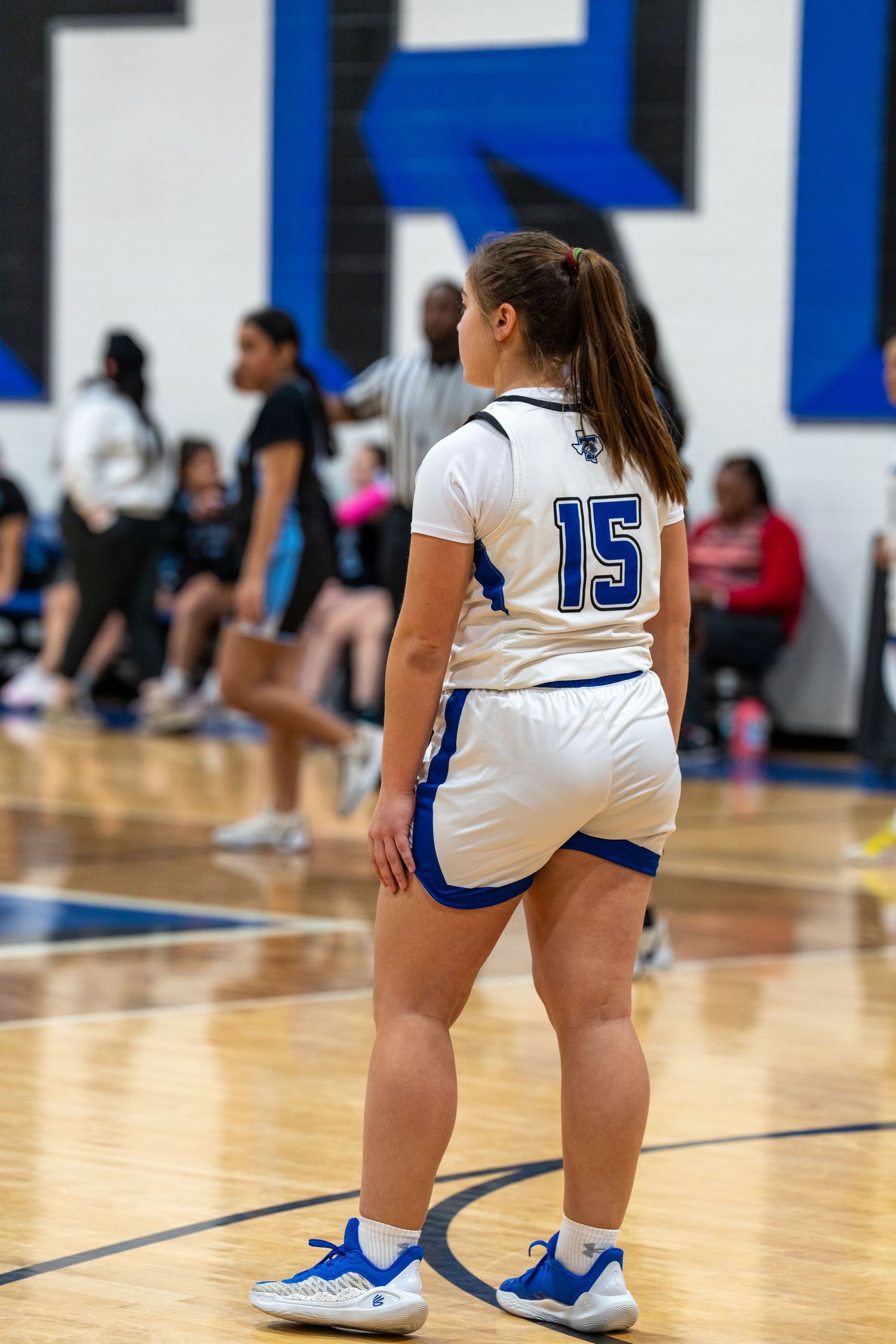 Volleyball players in white uniforms and blue shoes kneel on a shiny gymnasium floor during a match.