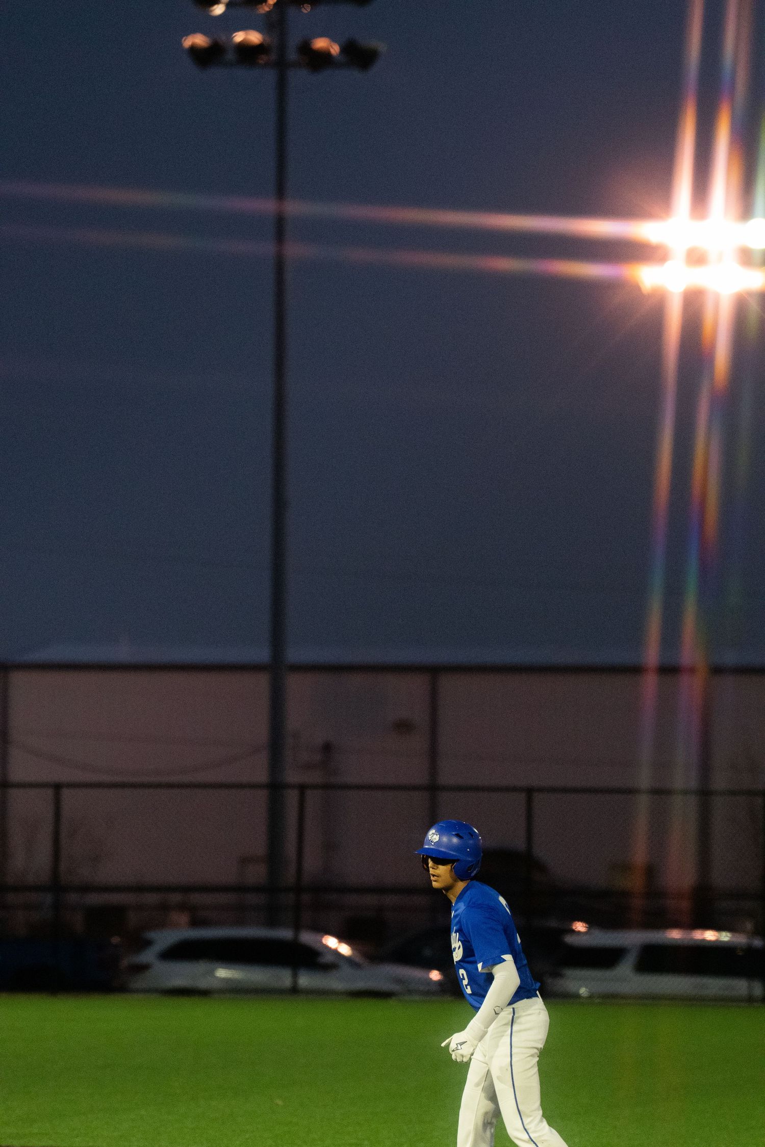 Baseball player in blue uniform throws a ball during twilight practice on green field with stadium lights glowing.