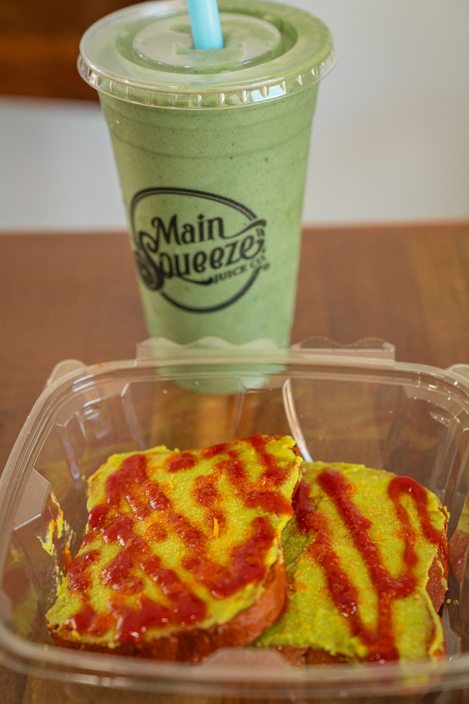 A green smoothie from Main Squeeze alongside two bright yellow sandwiches with red sauce drizzle in a clear container.