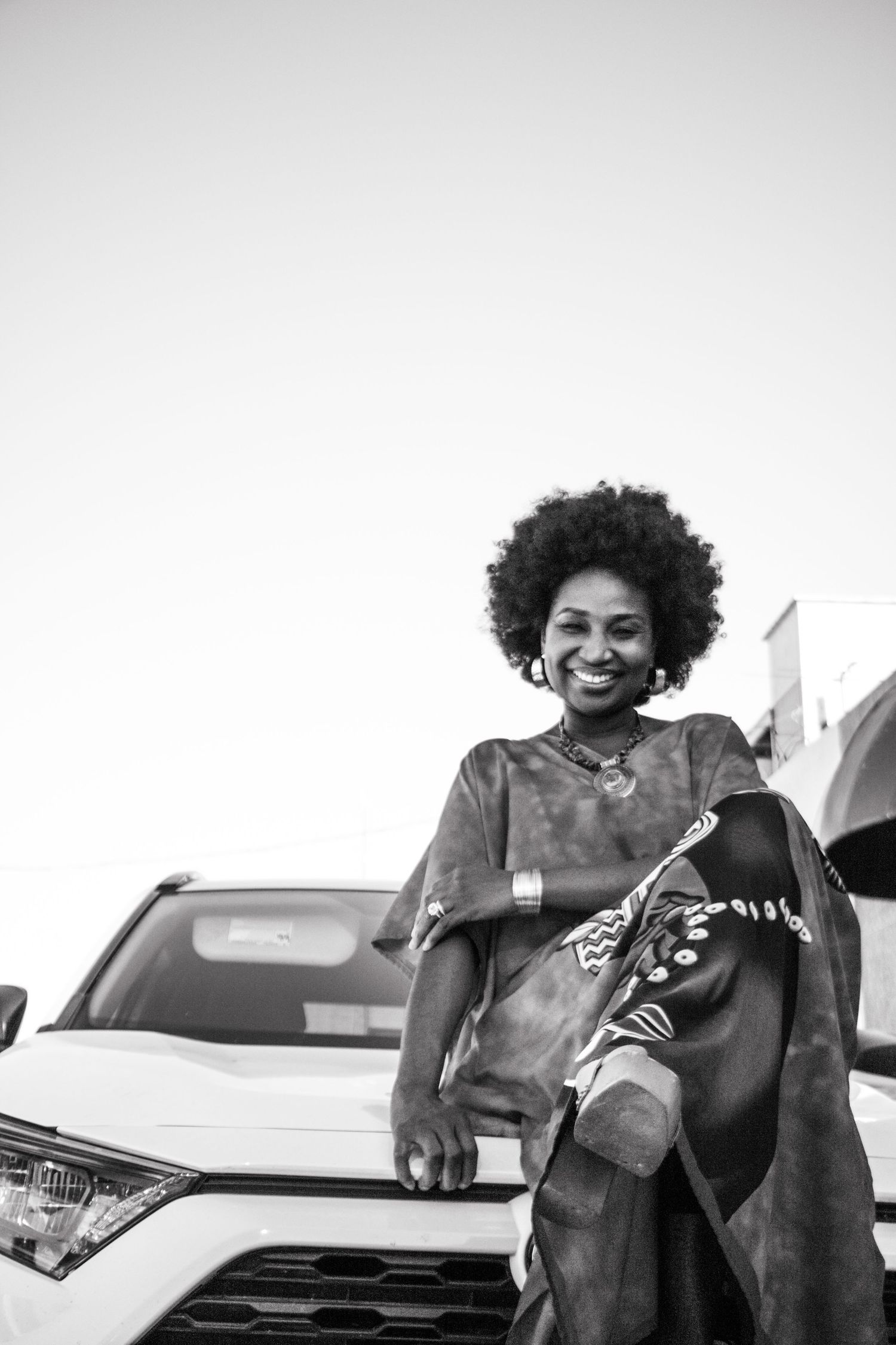 A stylish black and white portrait showing a person with natural hair posing confidently next to a white vehicle.