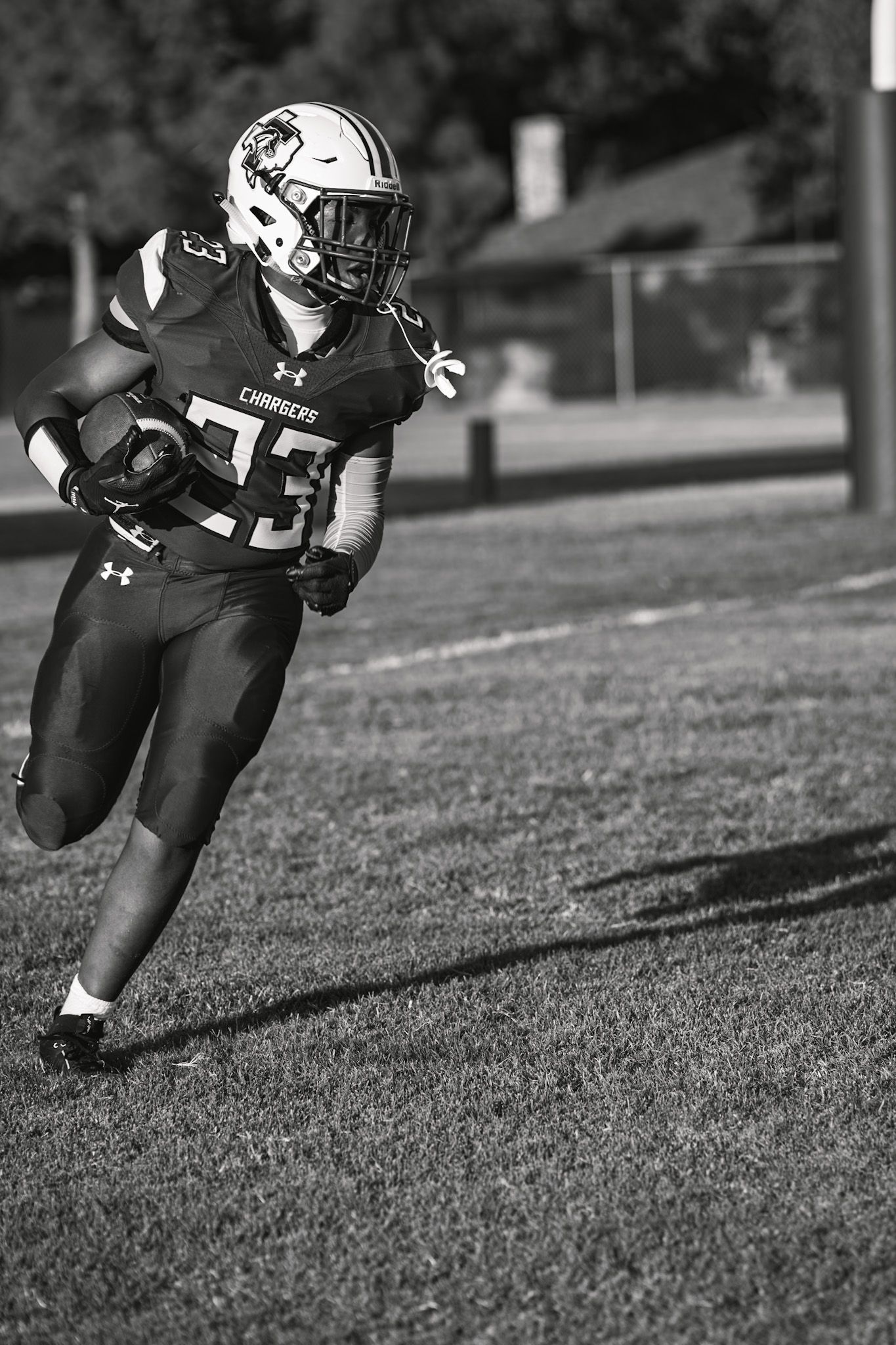 Black and white action photo of a football player in uniform running with the ball during a game on grass field.