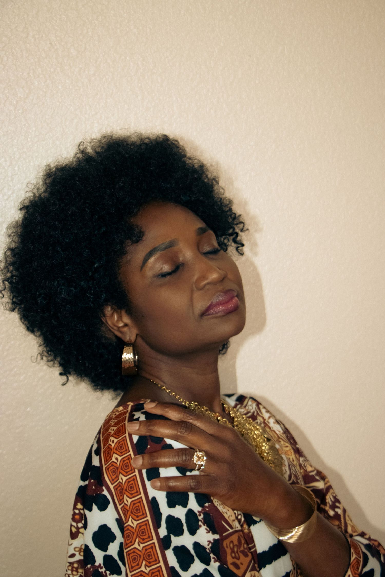 A serene portrait showing natural curly hair and jewelry against a neutral wall background.