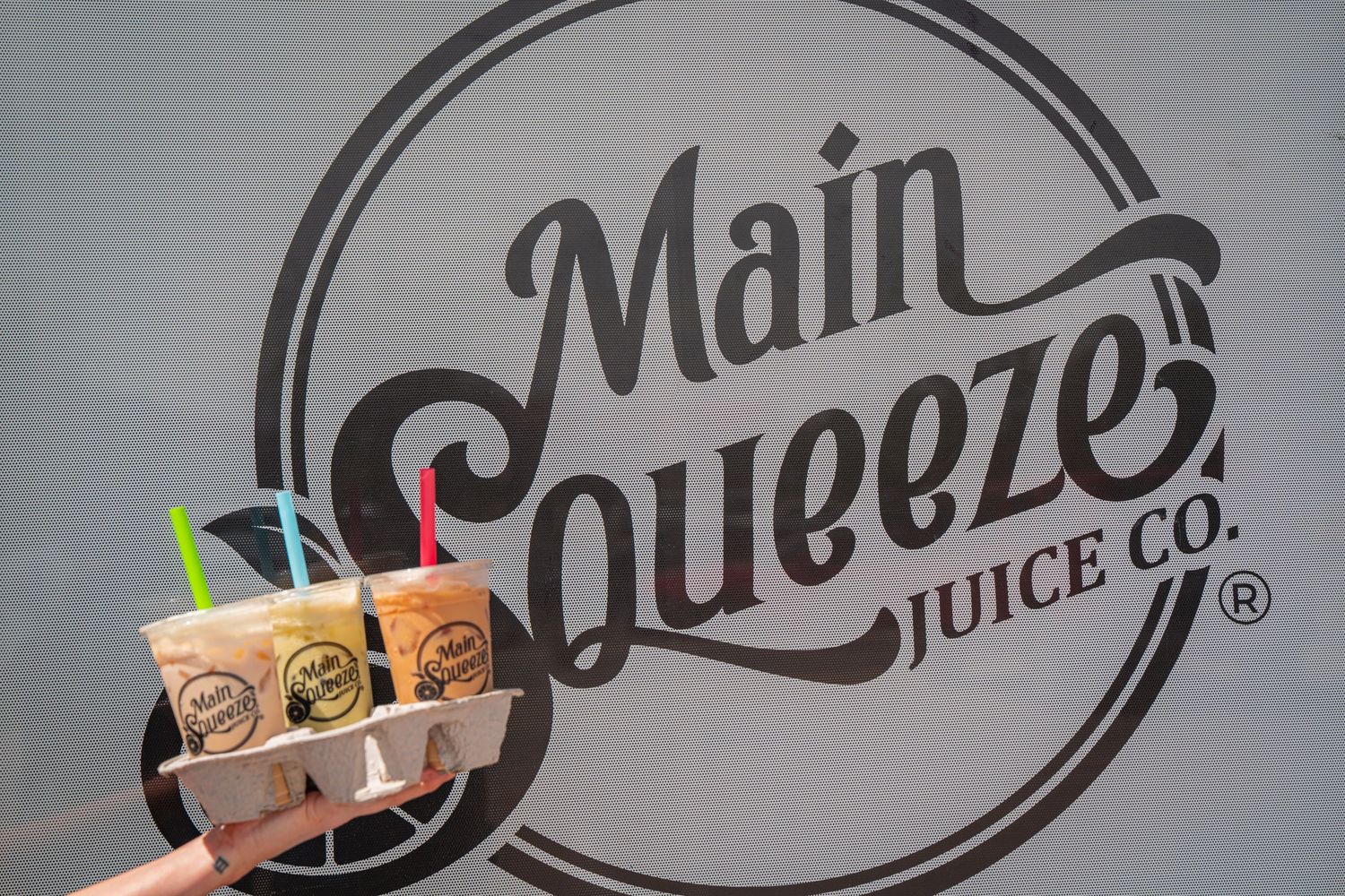 A hand holds colorful juice drinks in front of a circular Main Squeeze Juice Co. logo on a wall.