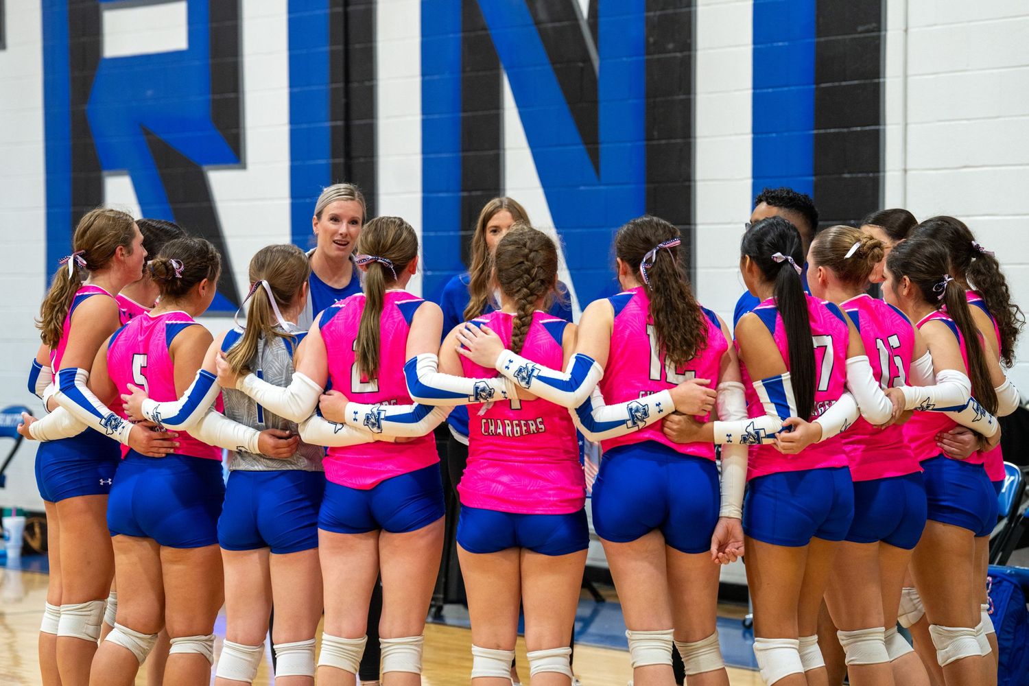Volleyball team huddles together wearing pink and blue uniforms during a game.