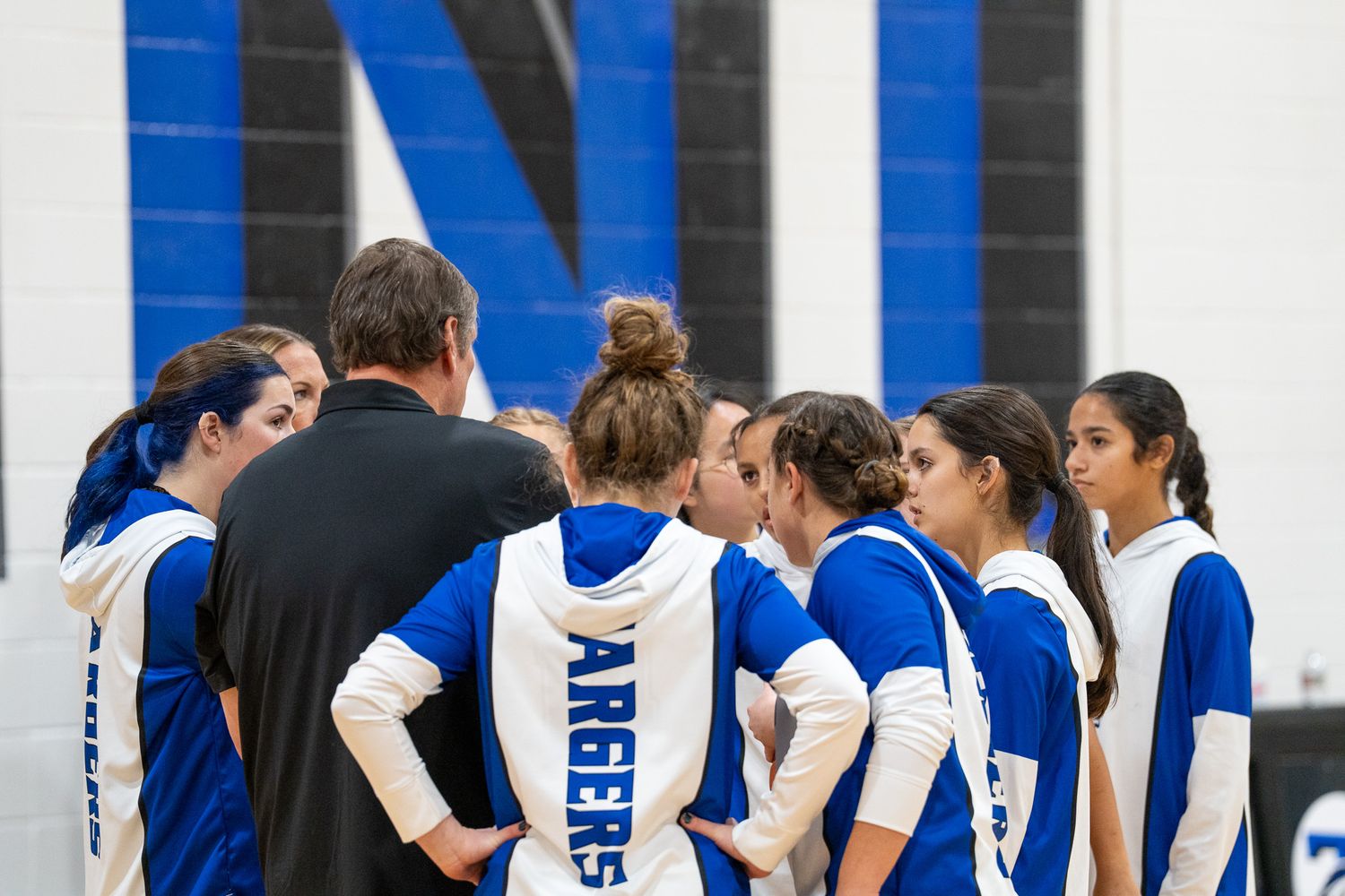 High school volleyball team in blue and white uniforms huddles together during a game break.