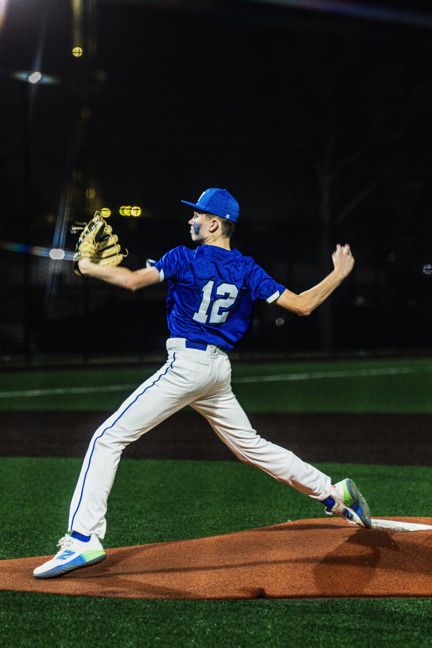 Baseball pitcher in blue jersey number 12 throws from the mound during a nighttime game.